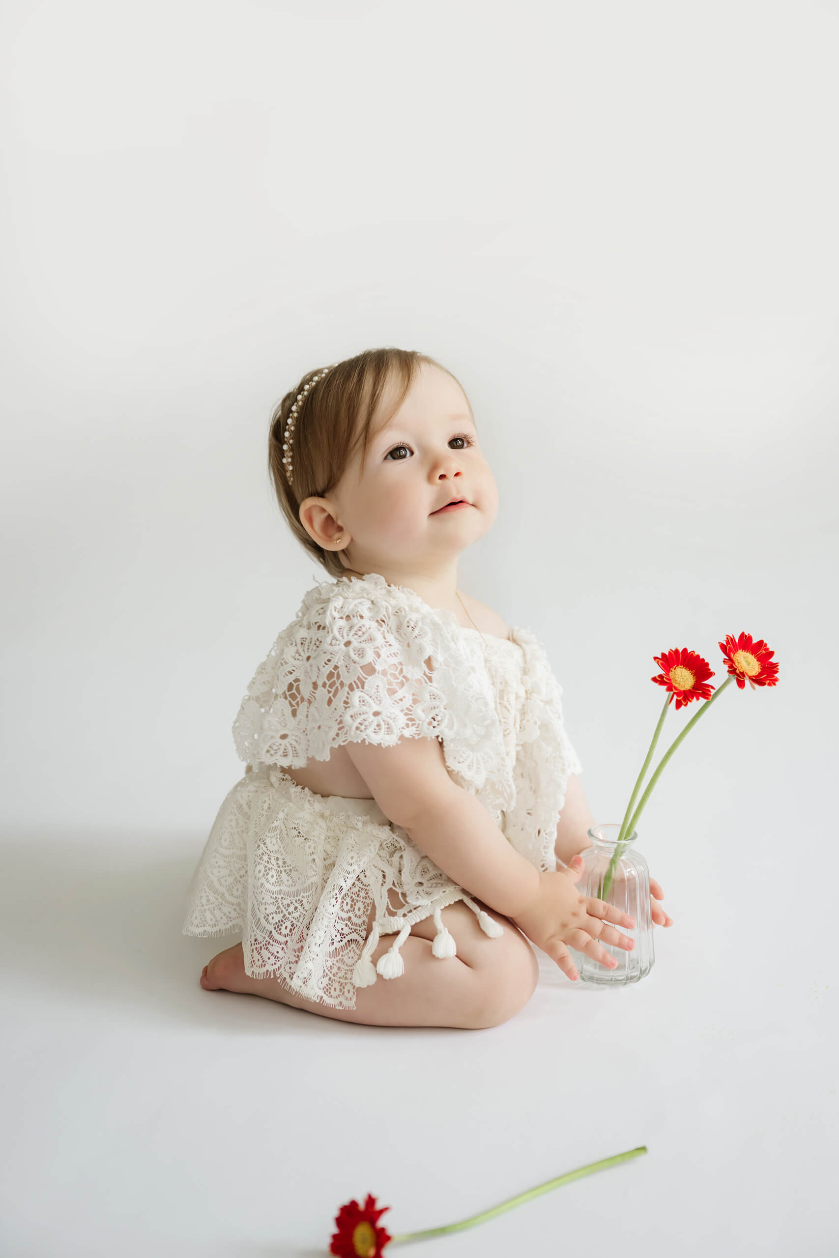 Baby girl kneeling with red flowers during a simple first birthday milestone photography session in a bright white studio.