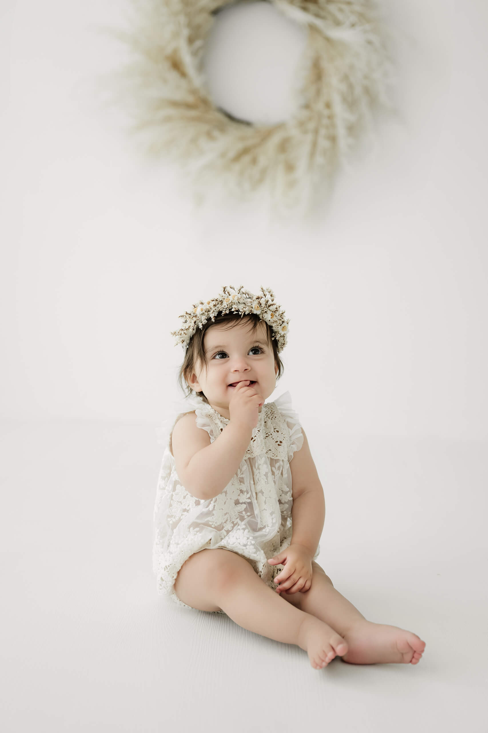 One-year-old girl wearing a floral crown during a simple first birthday milestone portrait session in a bright white studio.