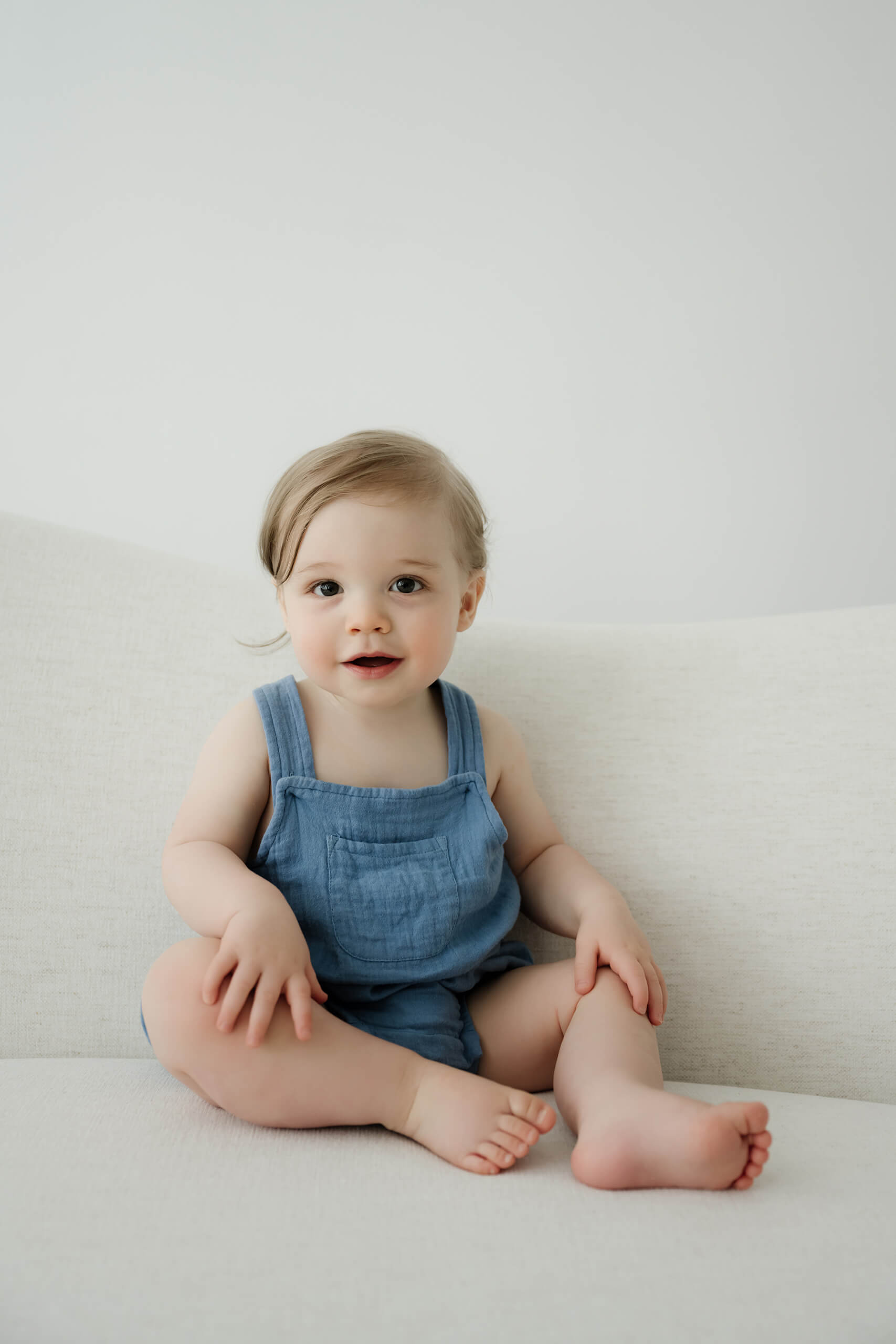 One-year-old sitting on a white studio sofa during a simple first birthday portrait session.