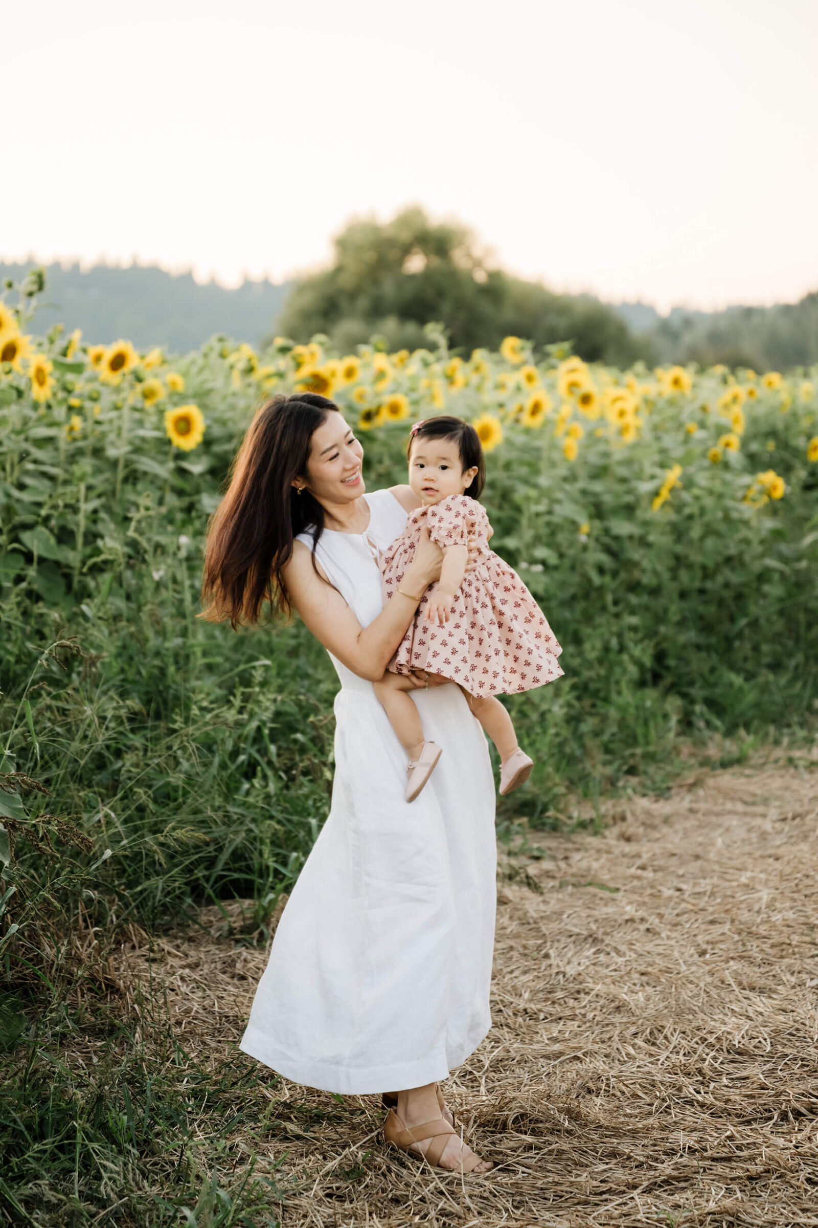 Mother holding her toddler daughter beside rows of sunflowers during a sunset family session in Duvall.