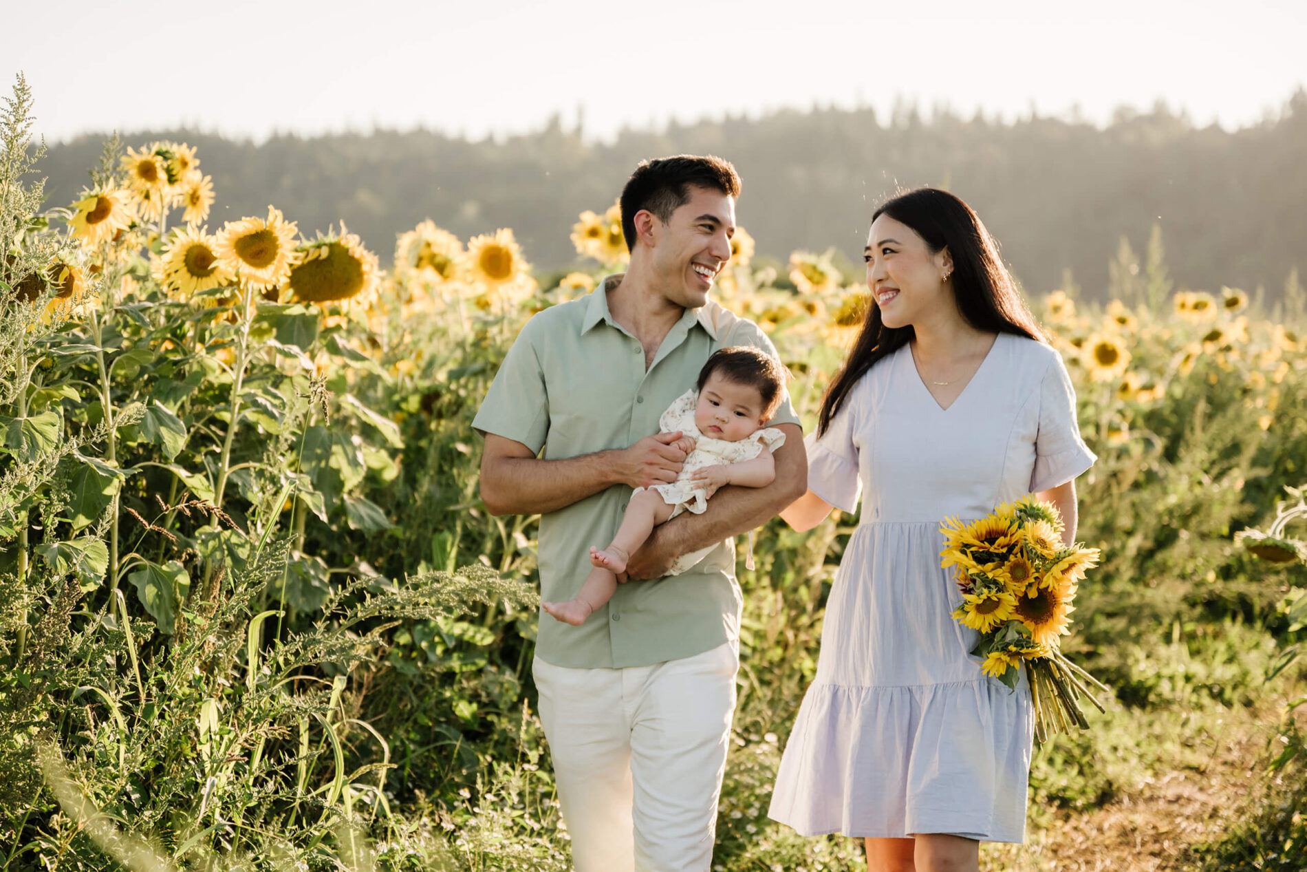Parents walking through a sunflower field with their baby and a bouquet during a summer family photo session near Seattle.