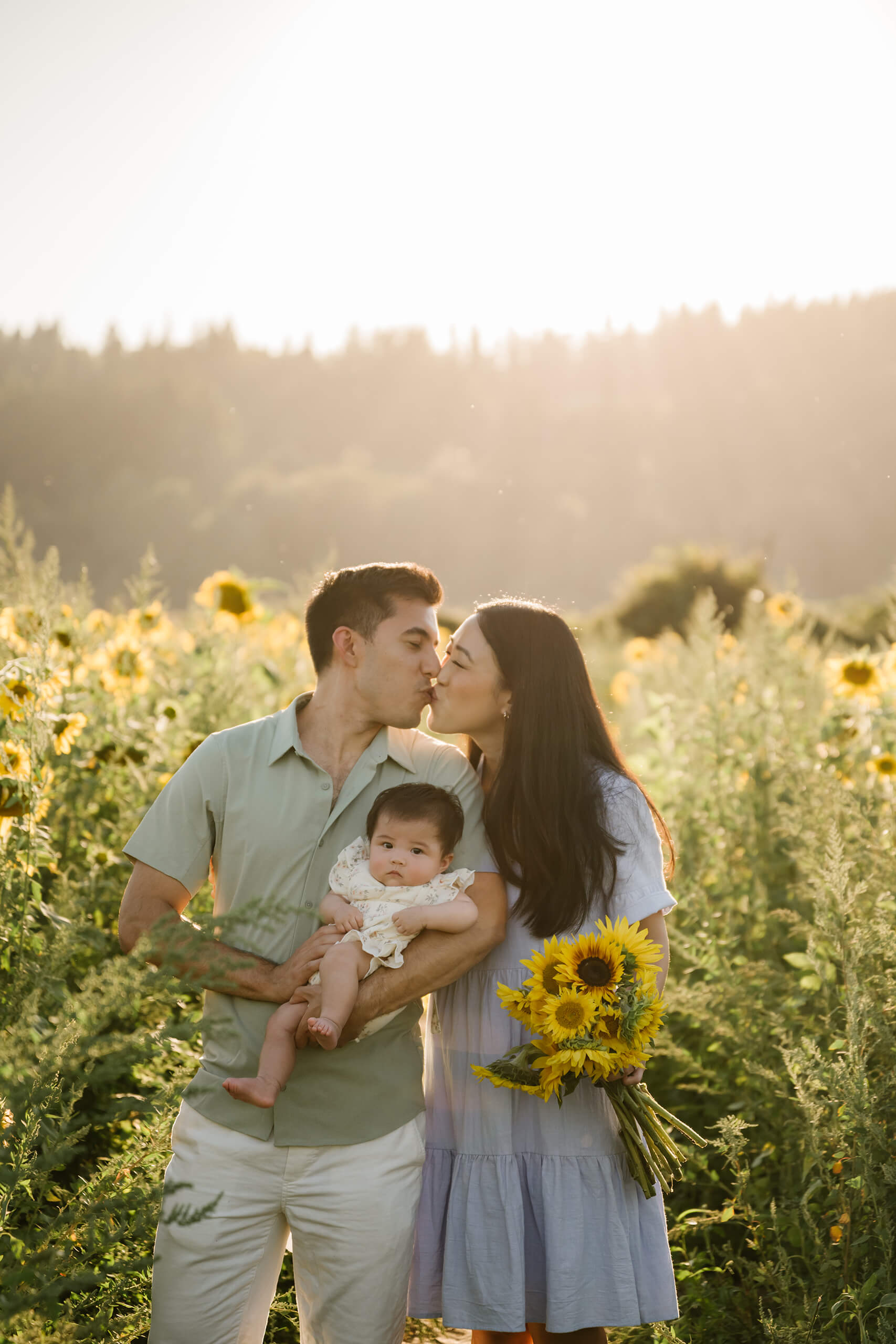 Parents kissing while holding their baby during a sunset sunflower field family session near Seattle.