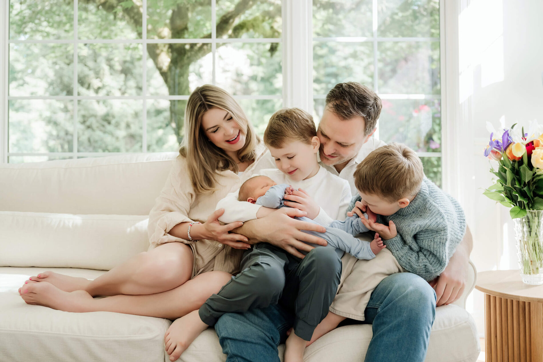Parents and two older brothers gather around their newborn baby on a sunlit sofa during an in-home newborn session.