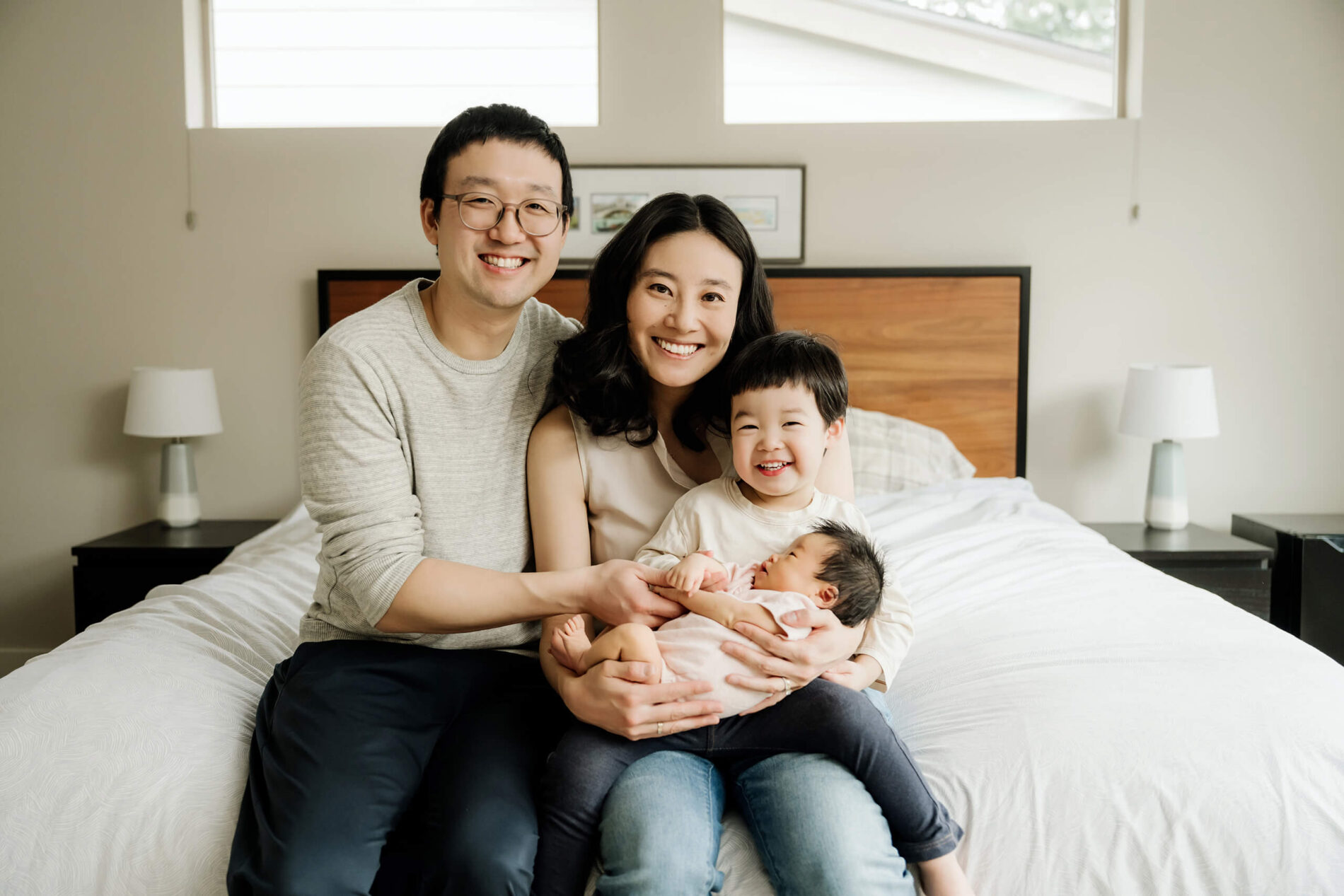 Family of four sits together on the bed while an older sibling holds the newborn baby in their lap.