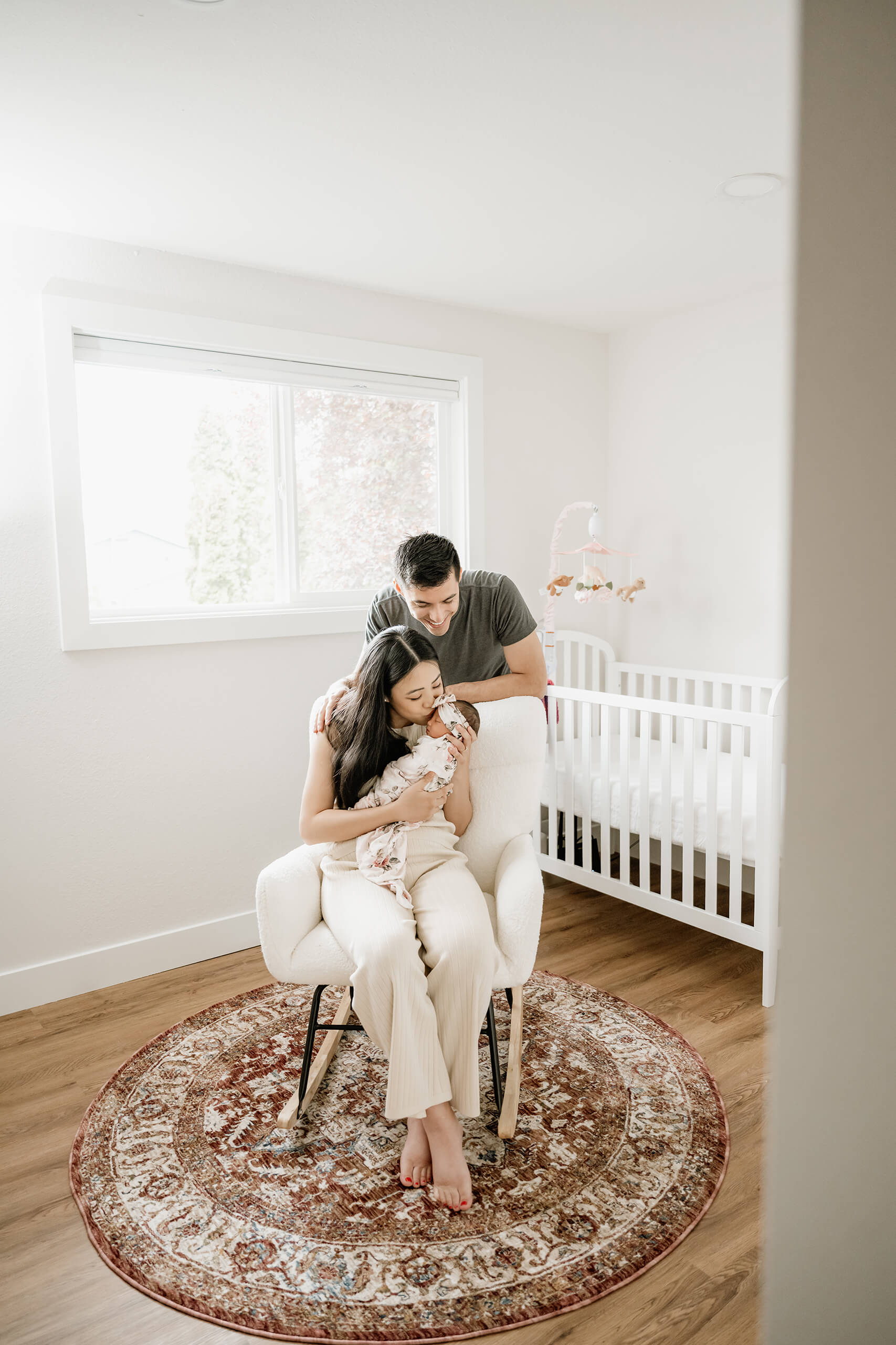 Mother holds and kisses her newborn baby in a nursery while the father stands beside her near the crib.