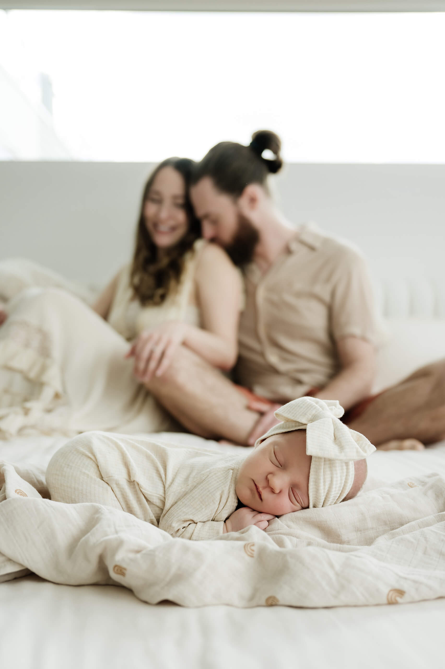 Sleeping newborn baby rests on a bed in the foreground while the parents sit together softly blurred behind her.