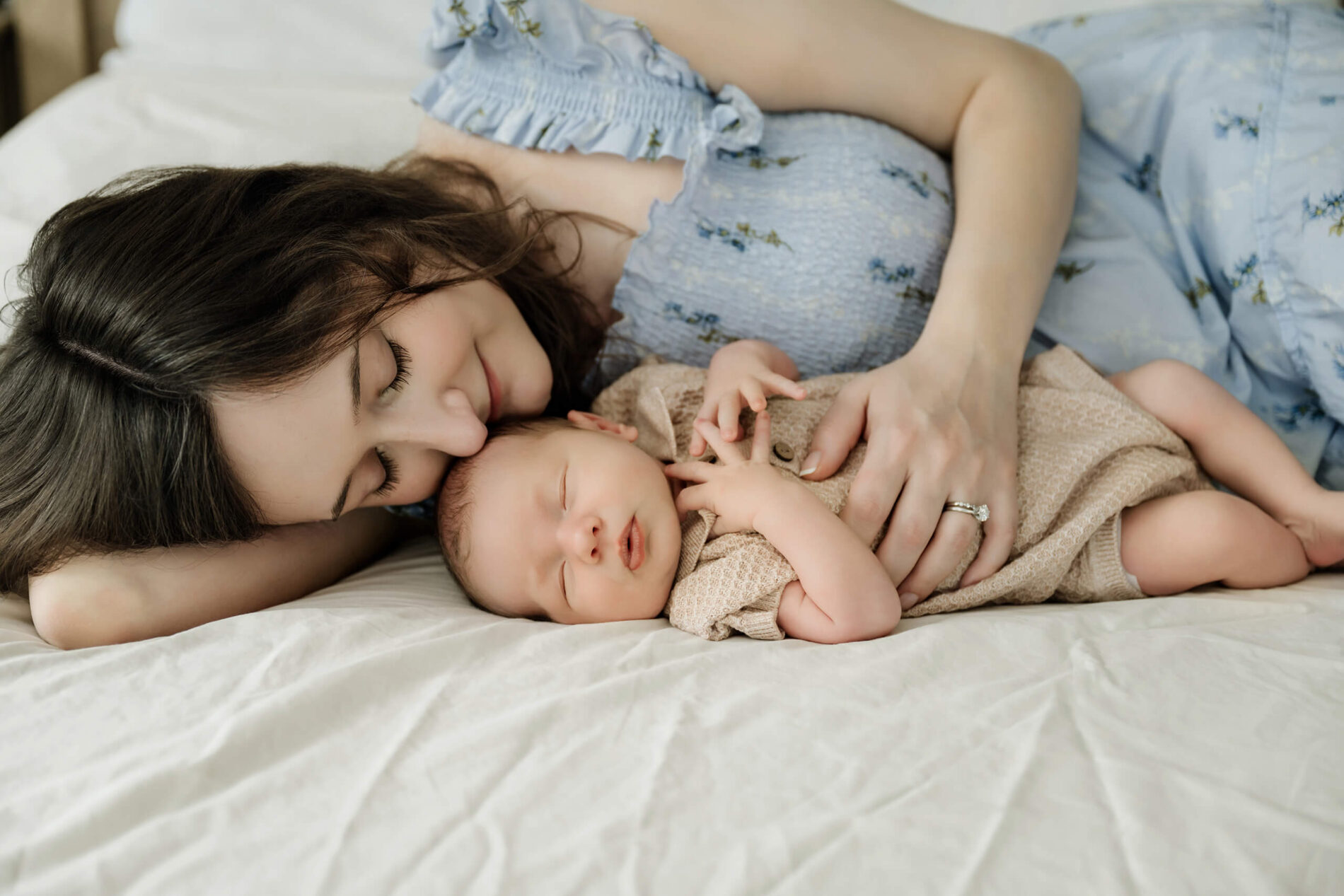 Mother lies beside her sleeping newborn baby and kisses the baby’s head during an in-home lifestyle session.