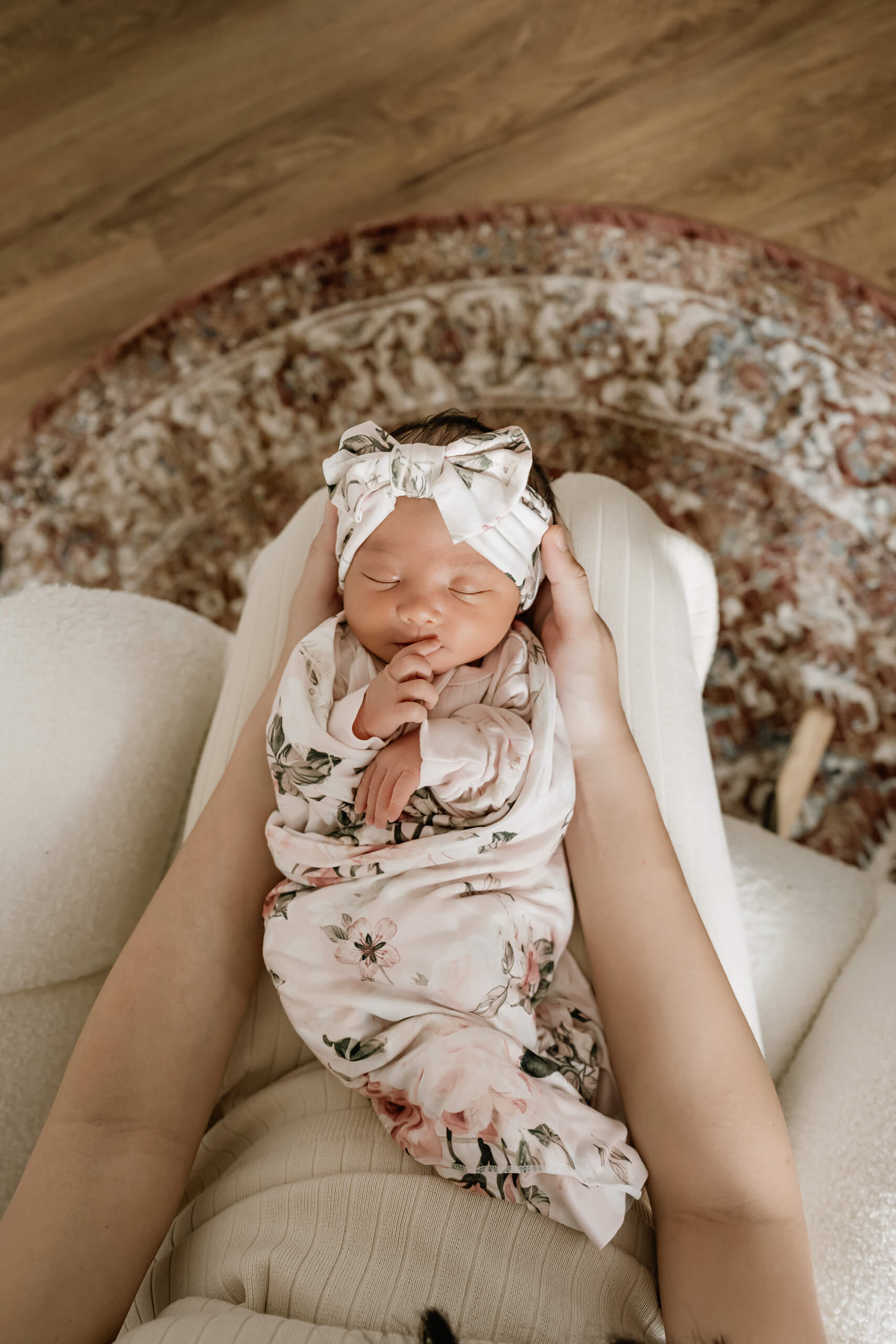 Newborn baby in a floral swaddle and matching head wrap rests in a parent’s lap above a patterned nursery rug.