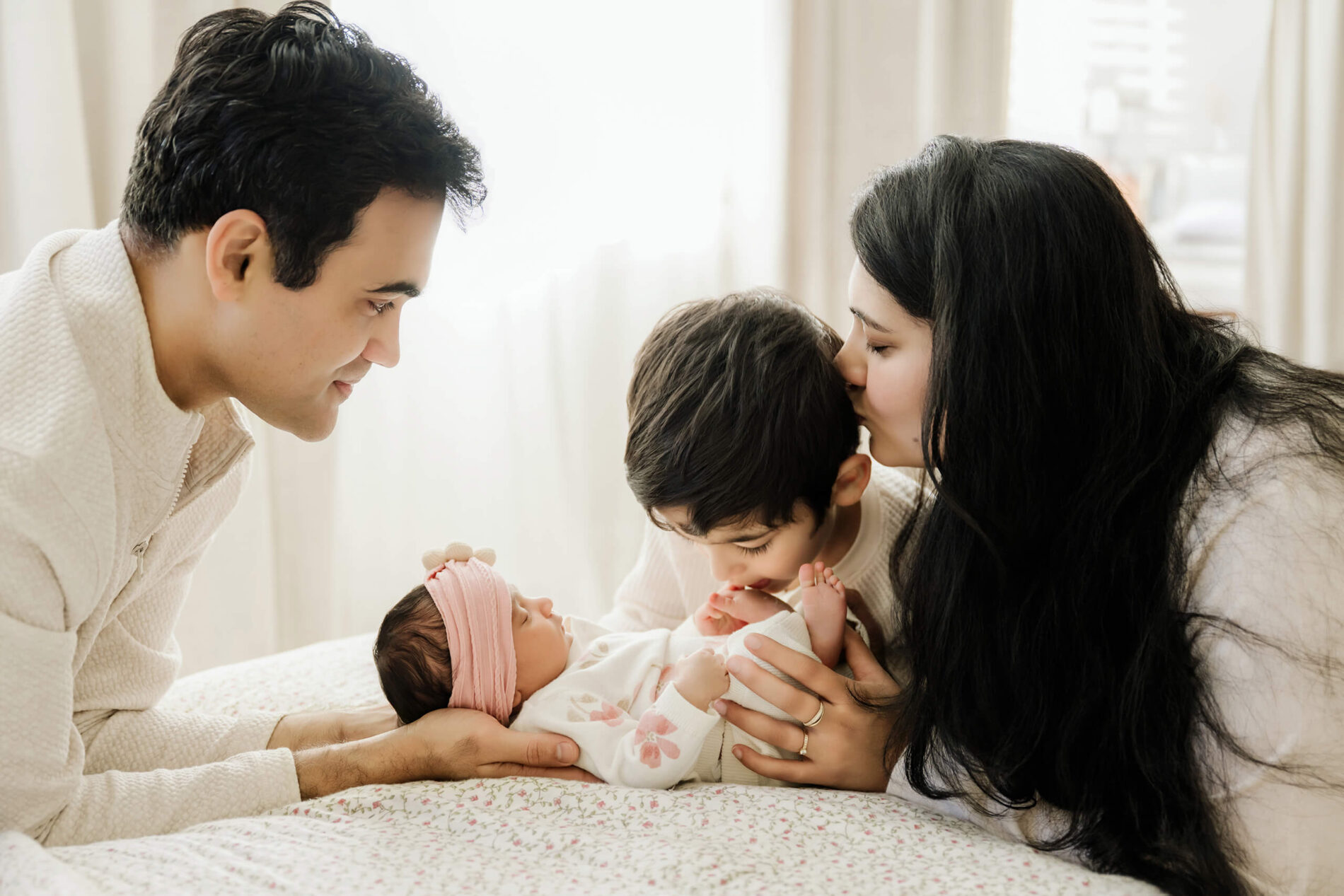 Parents and an older brother surround the newborn on a bed, sharing kisses and looking toward the baby.
