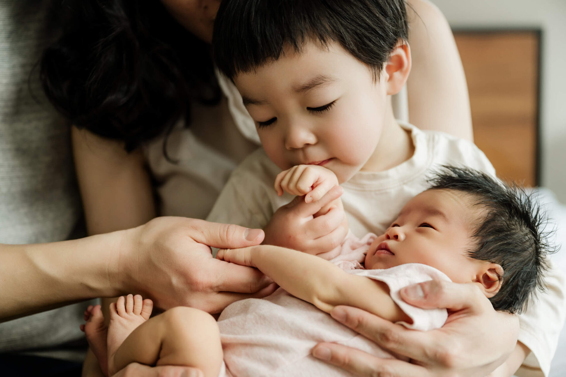 Older brother gently holds the newborn baby while the baby looks up at him, supported by their parents’ hands.