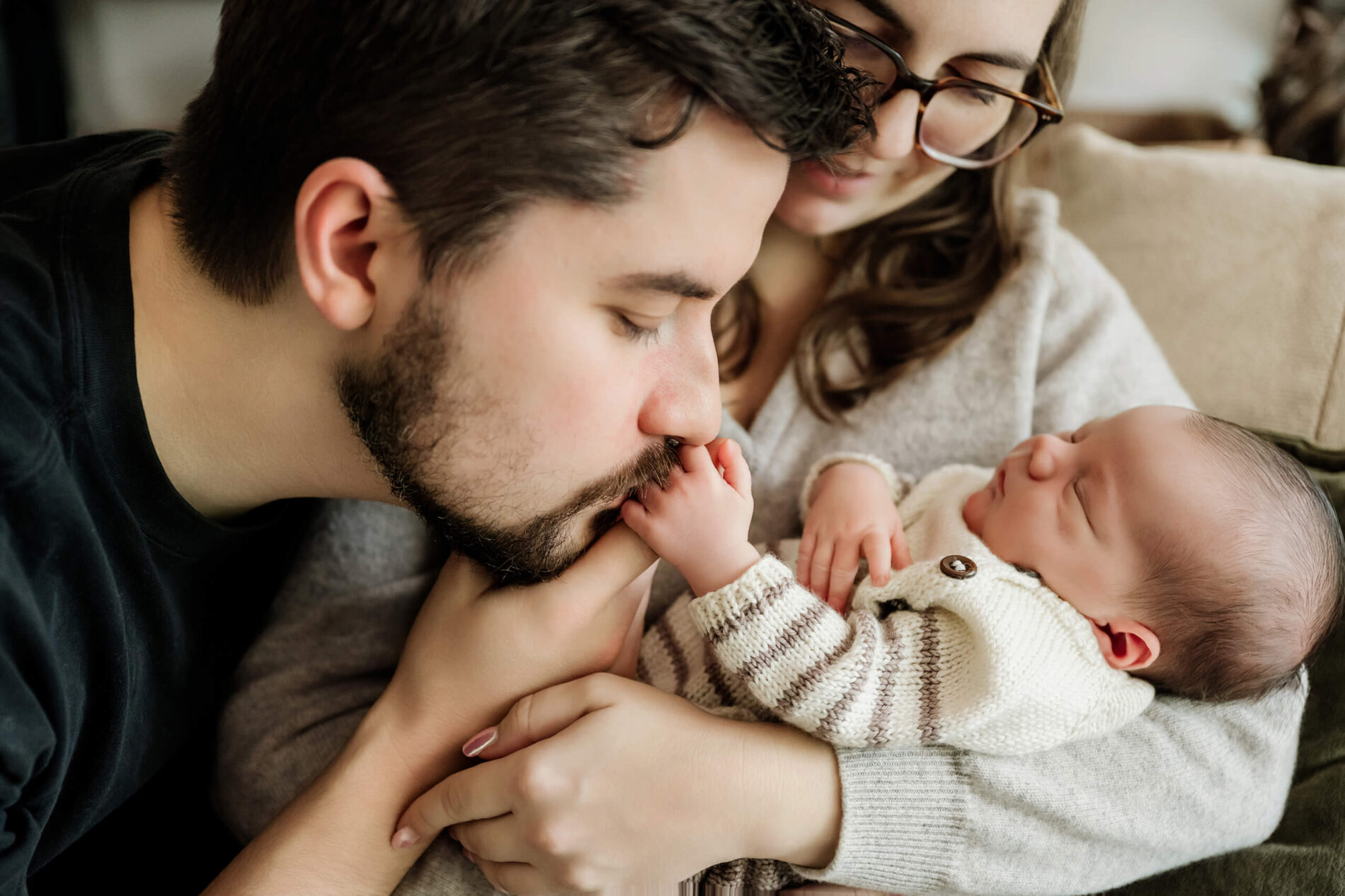 Father kisses his newborn baby’s foot while the mother cradles the sleeping baby in her arms at home.
