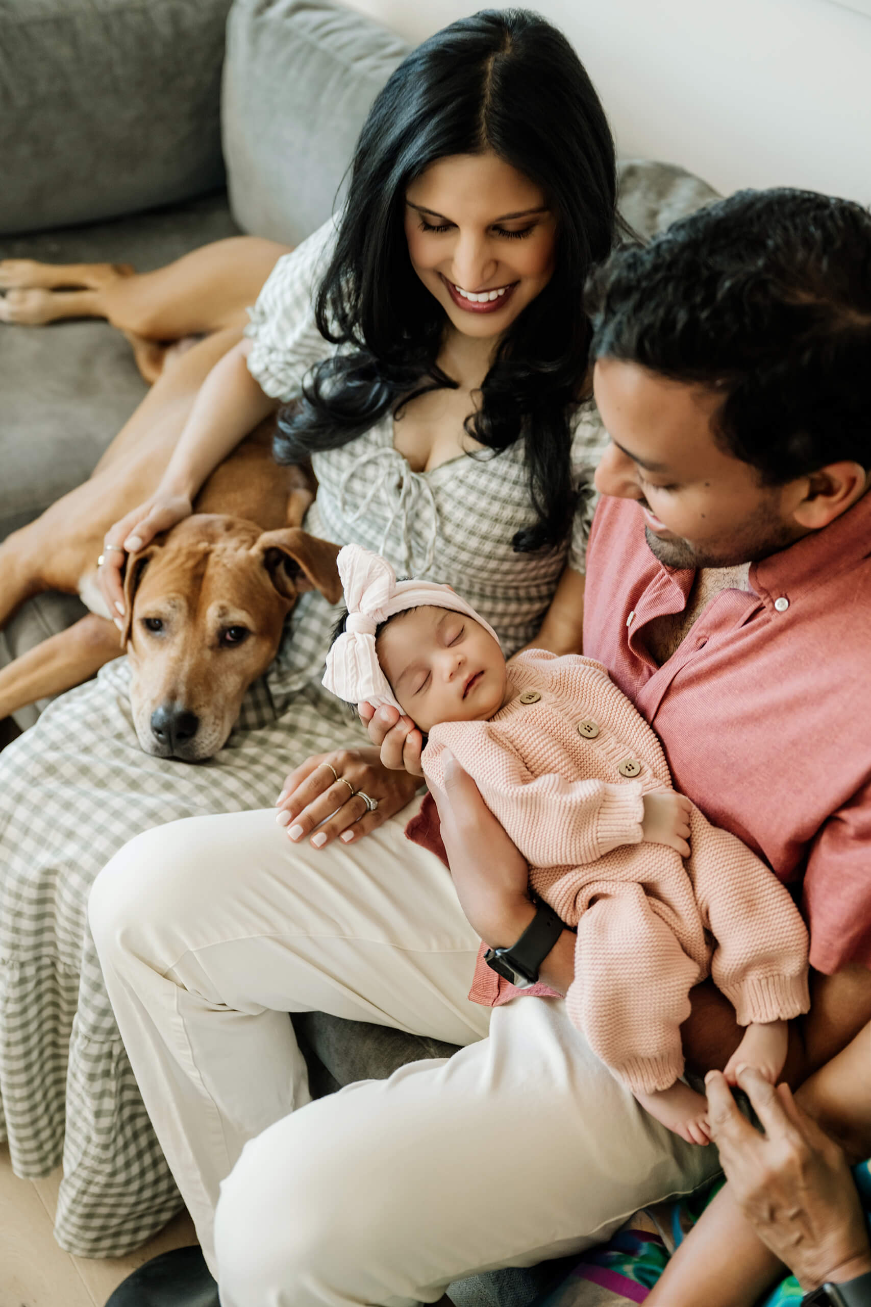 Parents sit together on a sofa with their sleeping newborn baby and the family dog during an in-home session.