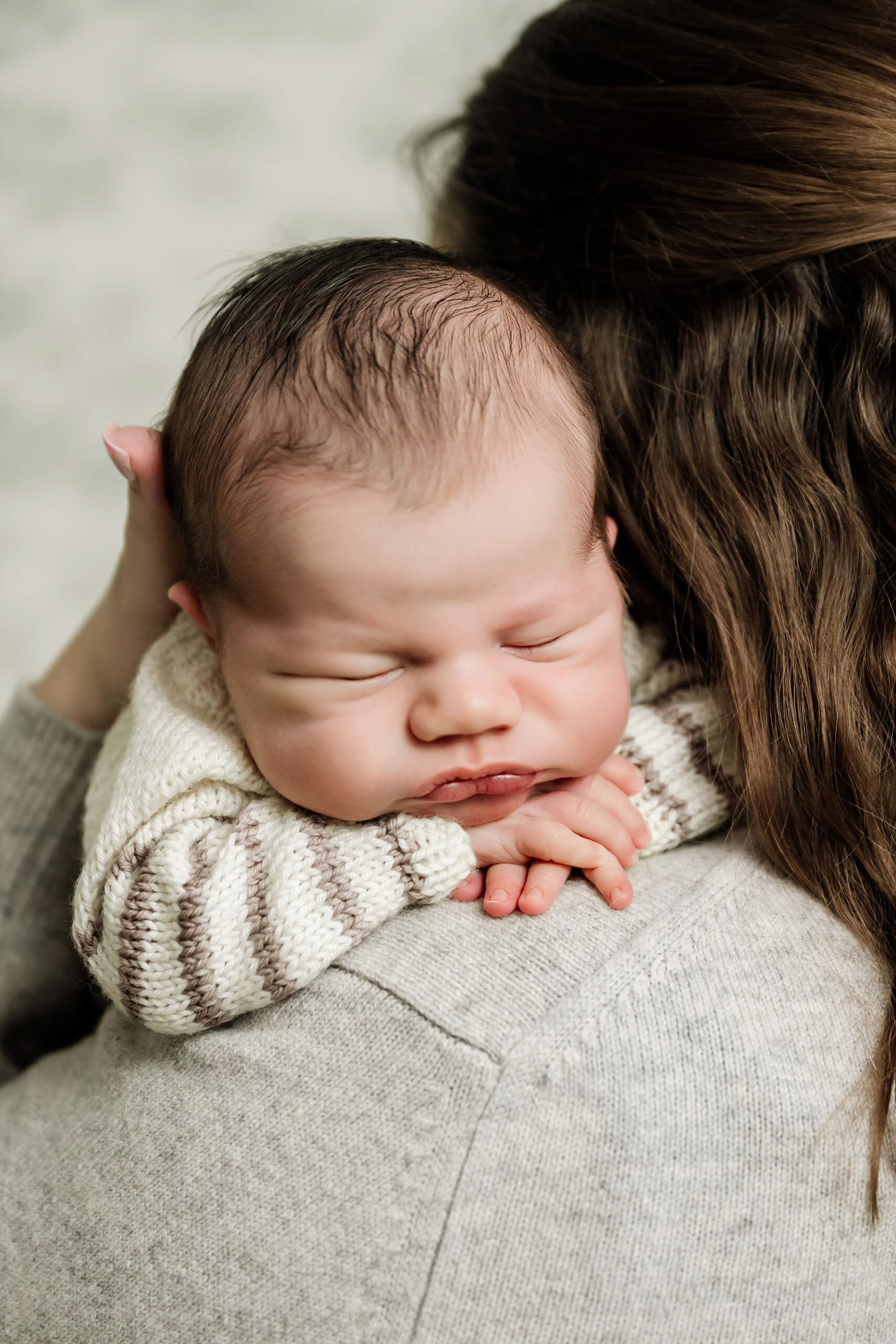 Sleeping newborn baby rests against the mother’s shoulder with folded hands beneath the chin.