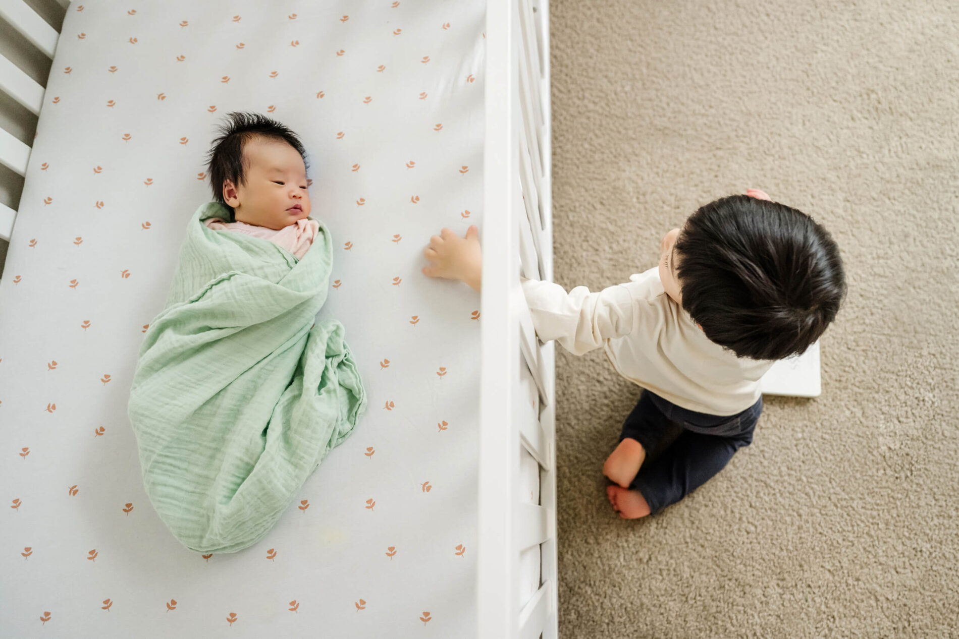 Older sibling kneels beside the crib and looks at the newborn baby wrapped in a light green blanket.