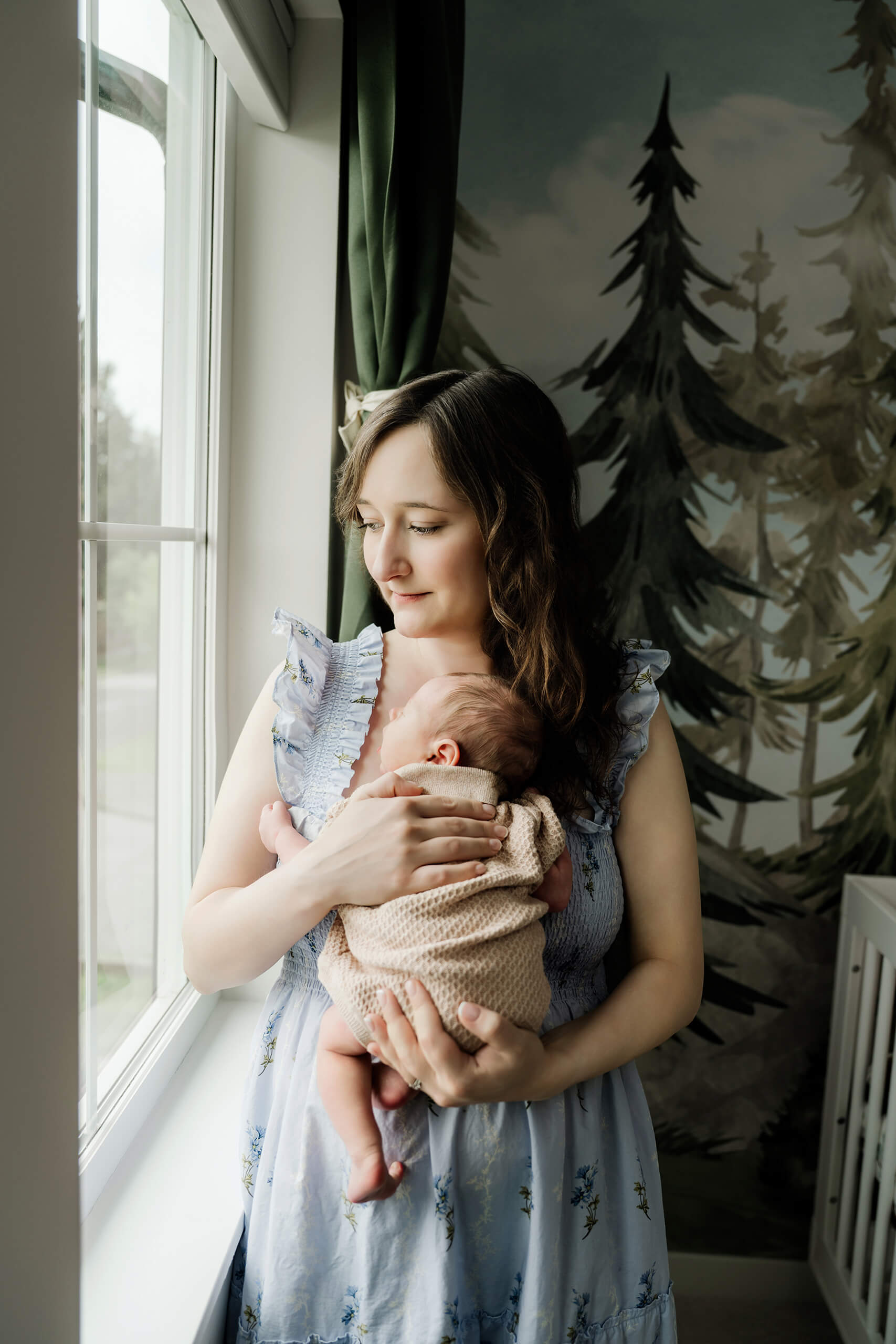 Mother holds her newborn baby beside a nursery window with a painted forest mural behind them.