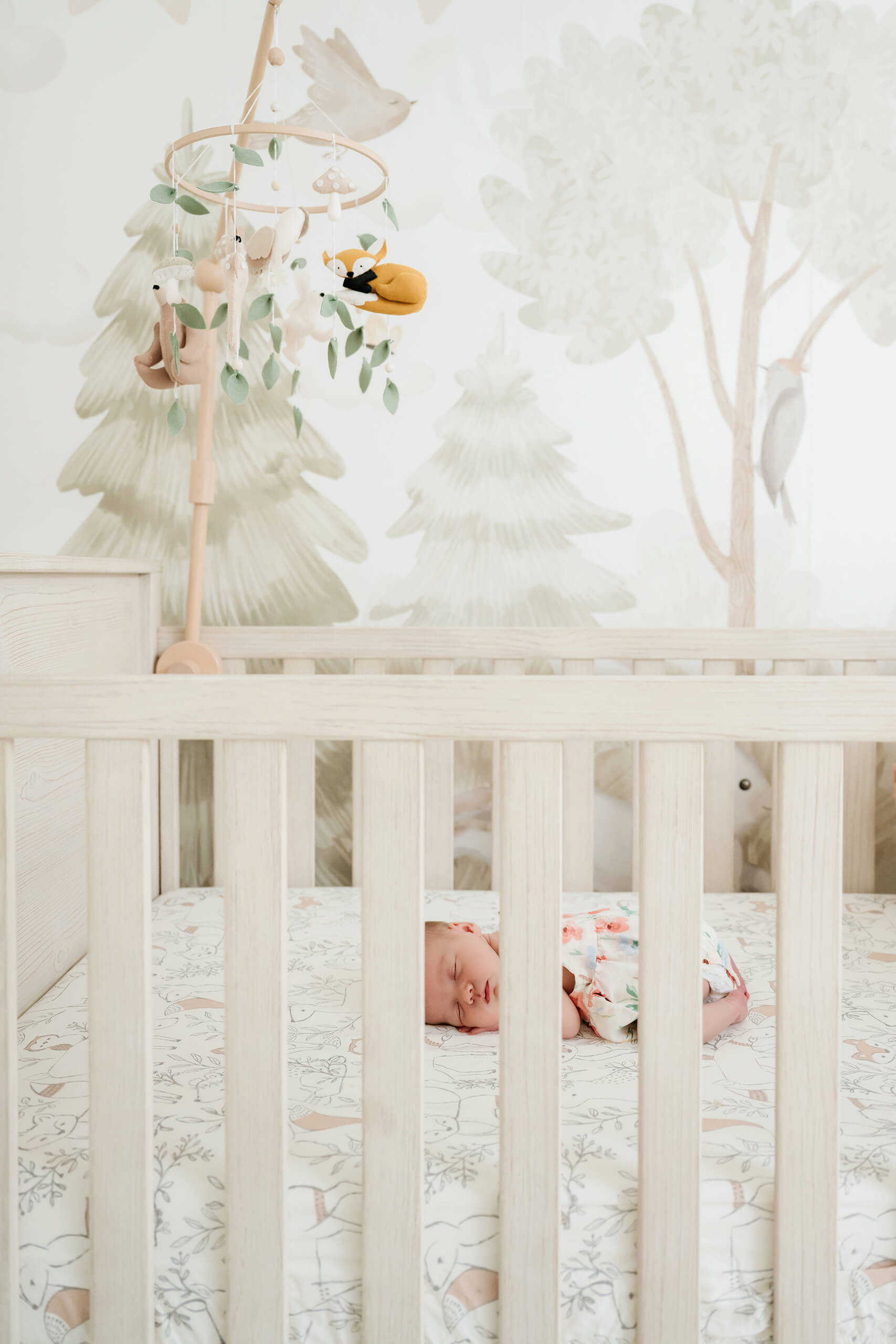 Newborn baby sleeps in a white crib beneath a woodland mobile in a softly decorated nursery.