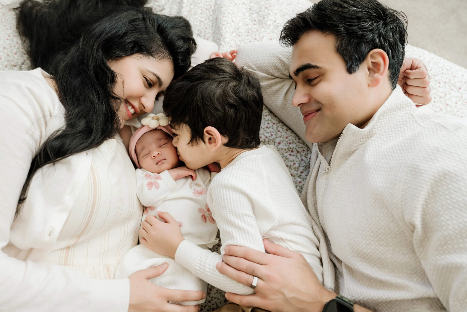 Parents and an older brother lie together on a bed while the brother kisses the sleeping newborn baby.