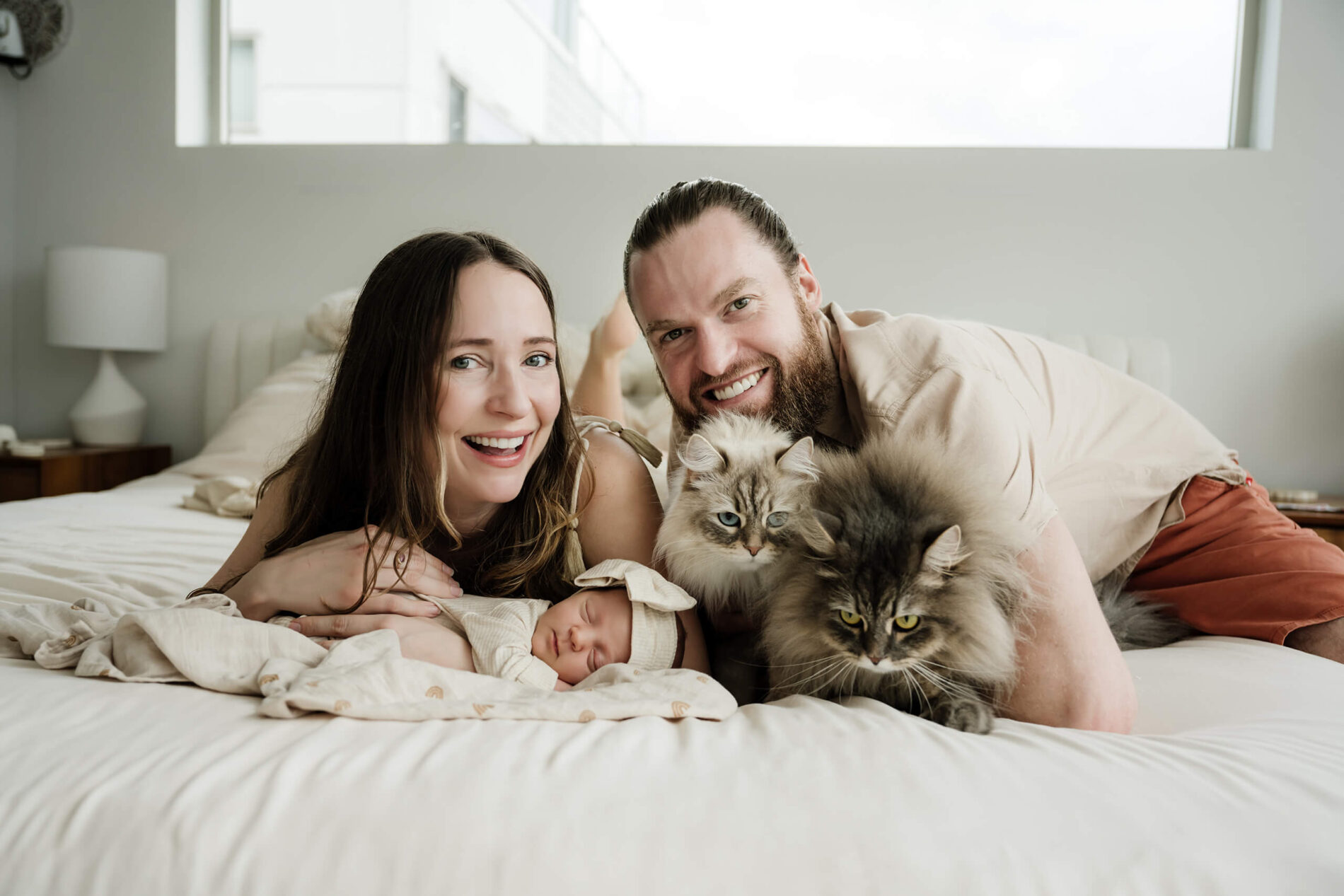 Parents lie on a bed with their sleeping newborn baby and two long-haired family cats.