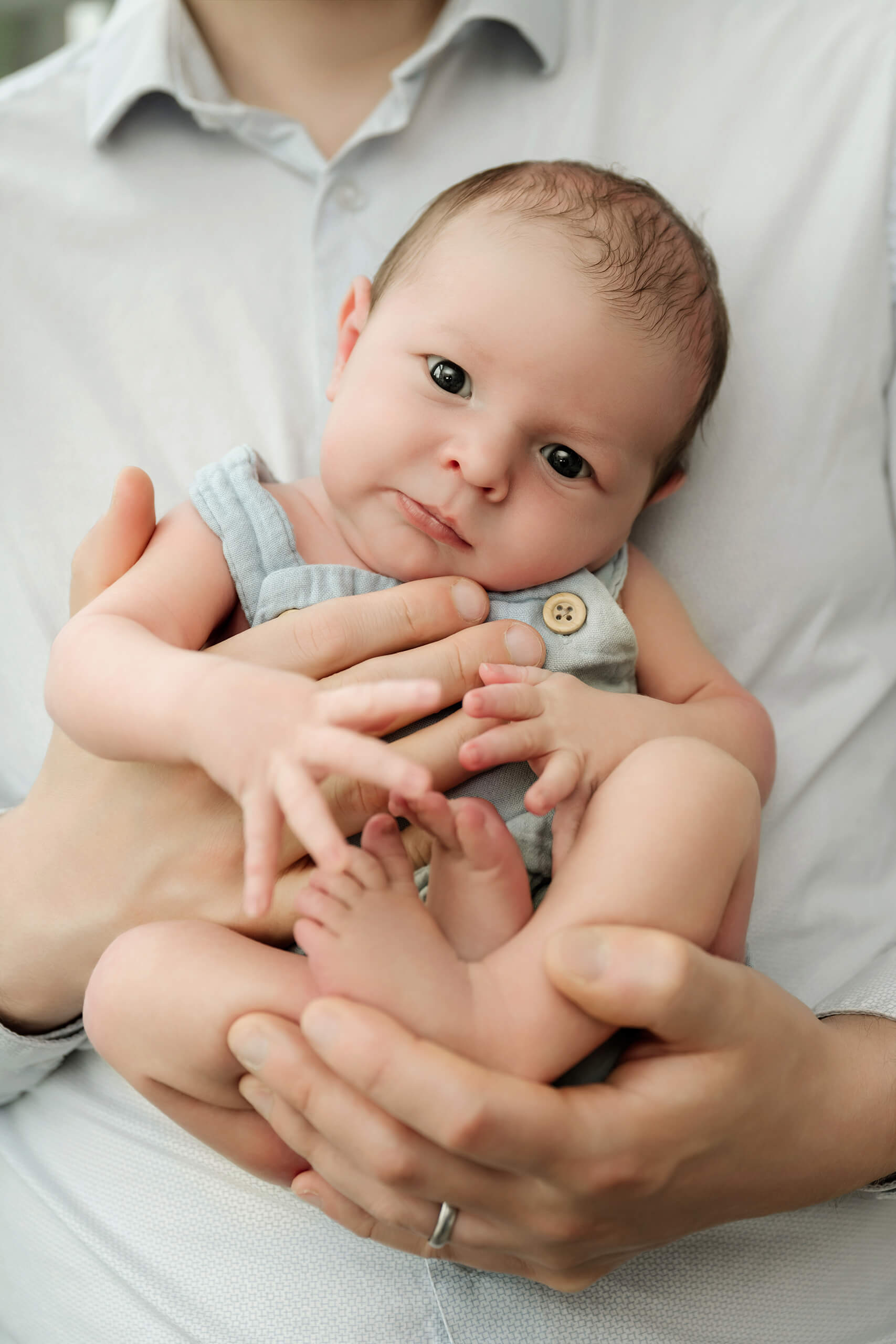 Awake newborn baby in a light blue romper rests securely in the father’s hands.