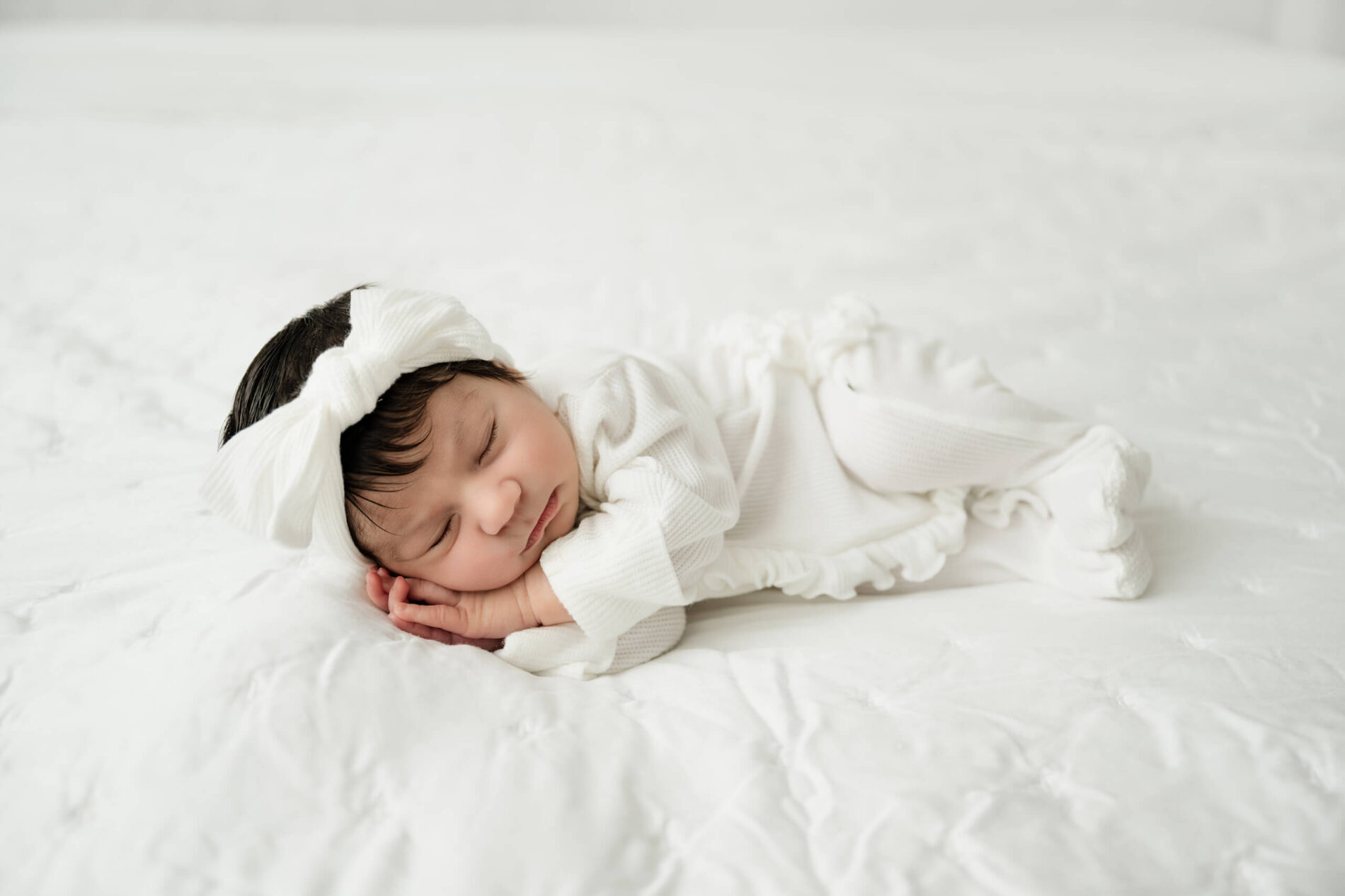 Sleeping newborn baby wears a white outfit and oversized bow while resting on a white bed.
