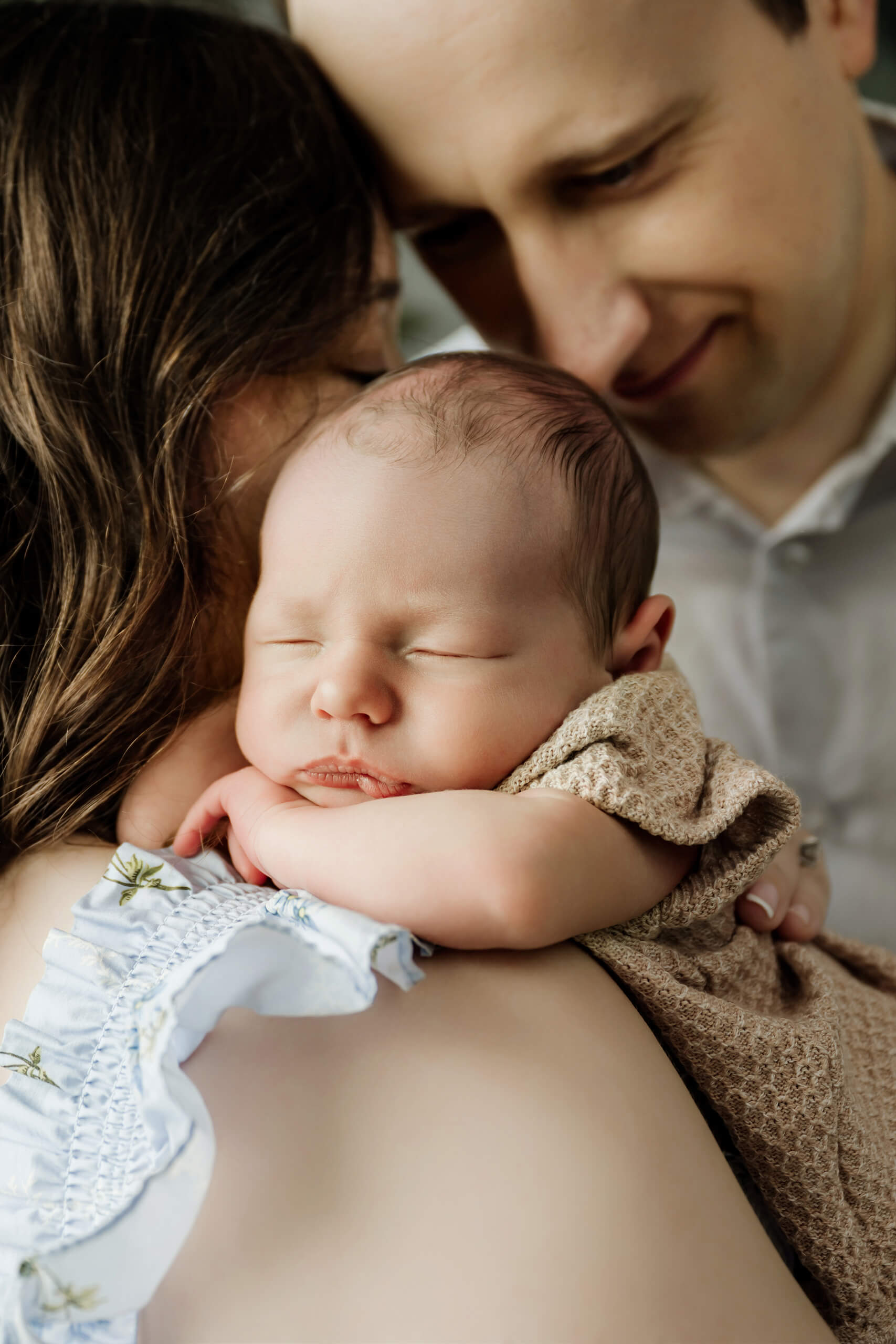 Sleeping newborn baby rests over the mother’s shoulder with the father close beside them.