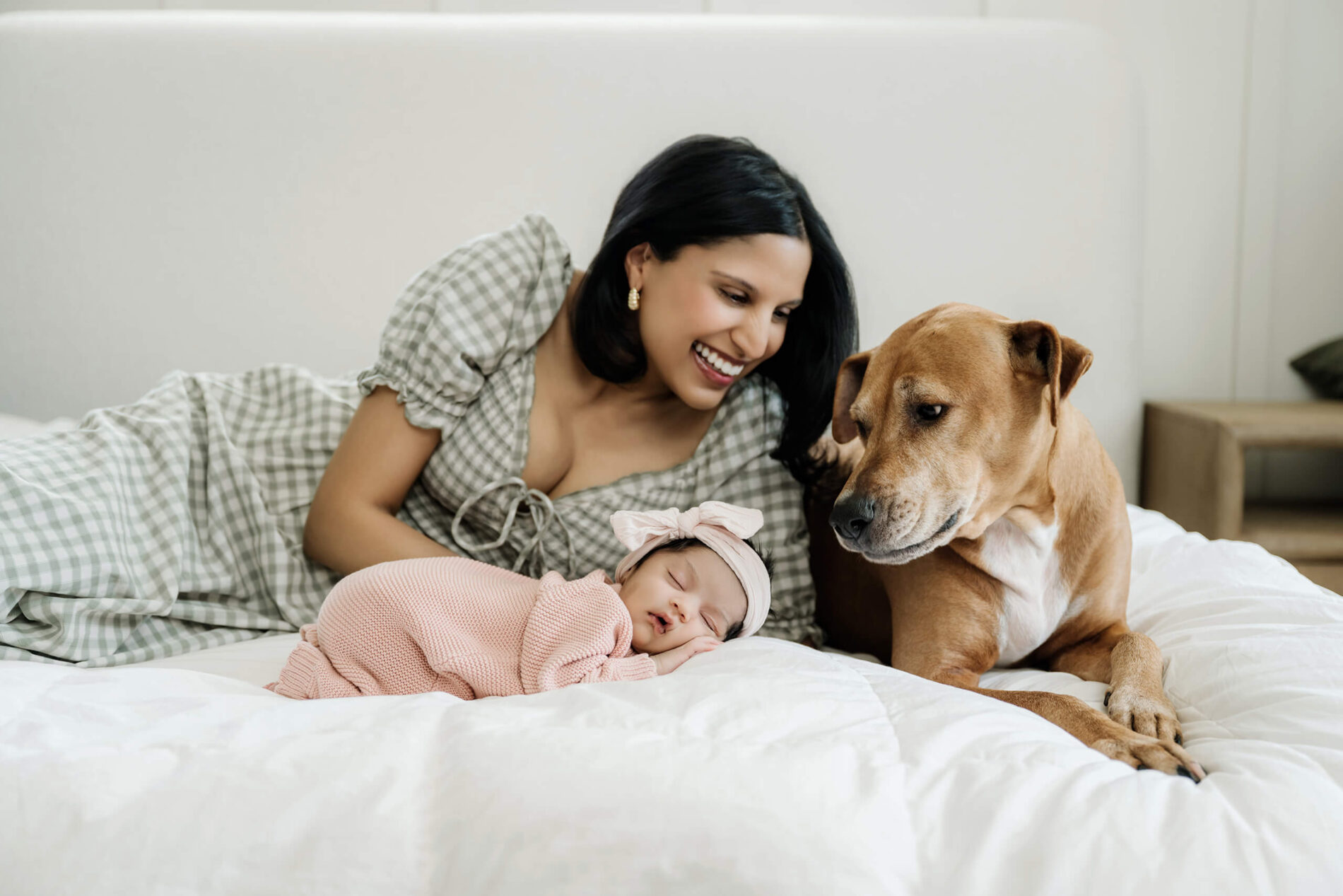 Mother smiles at her sleeping newborn baby beside the family dog during an in-home session.