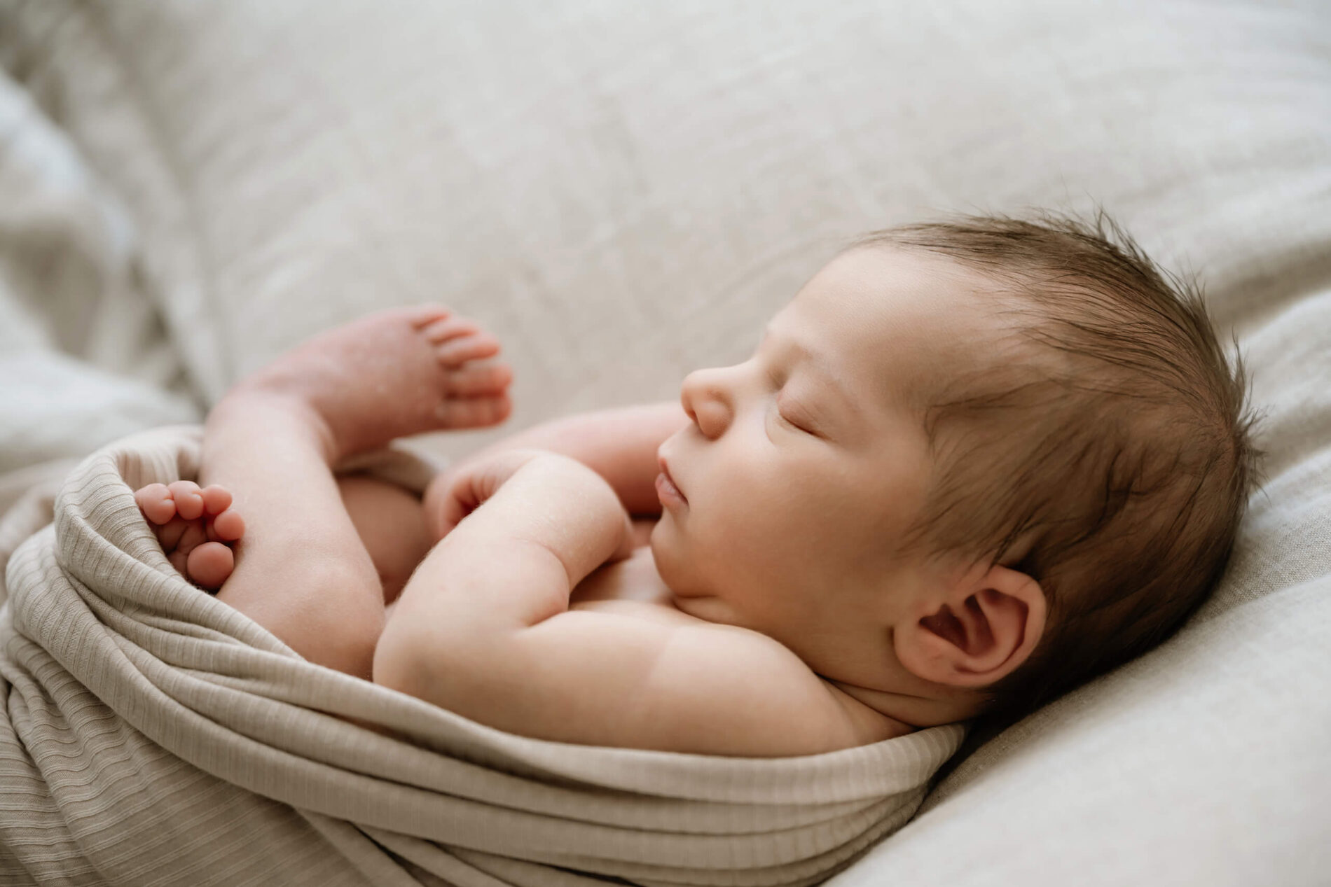 Sleeping newborn baby rests naturally on a bed with a soft neutral wrap draped around the body.