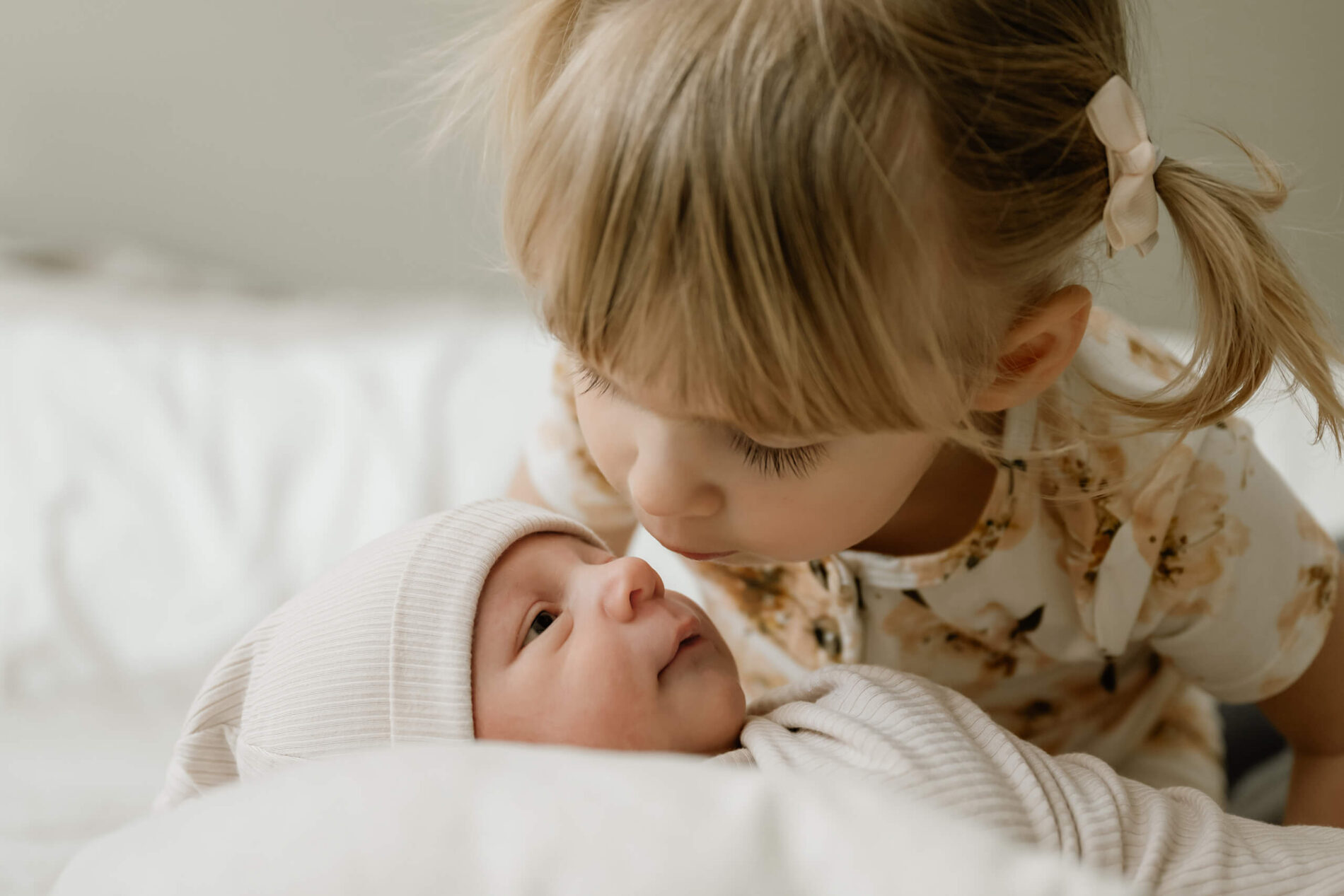 Older sister leans in to kiss her awake newborn sibling on the bed.