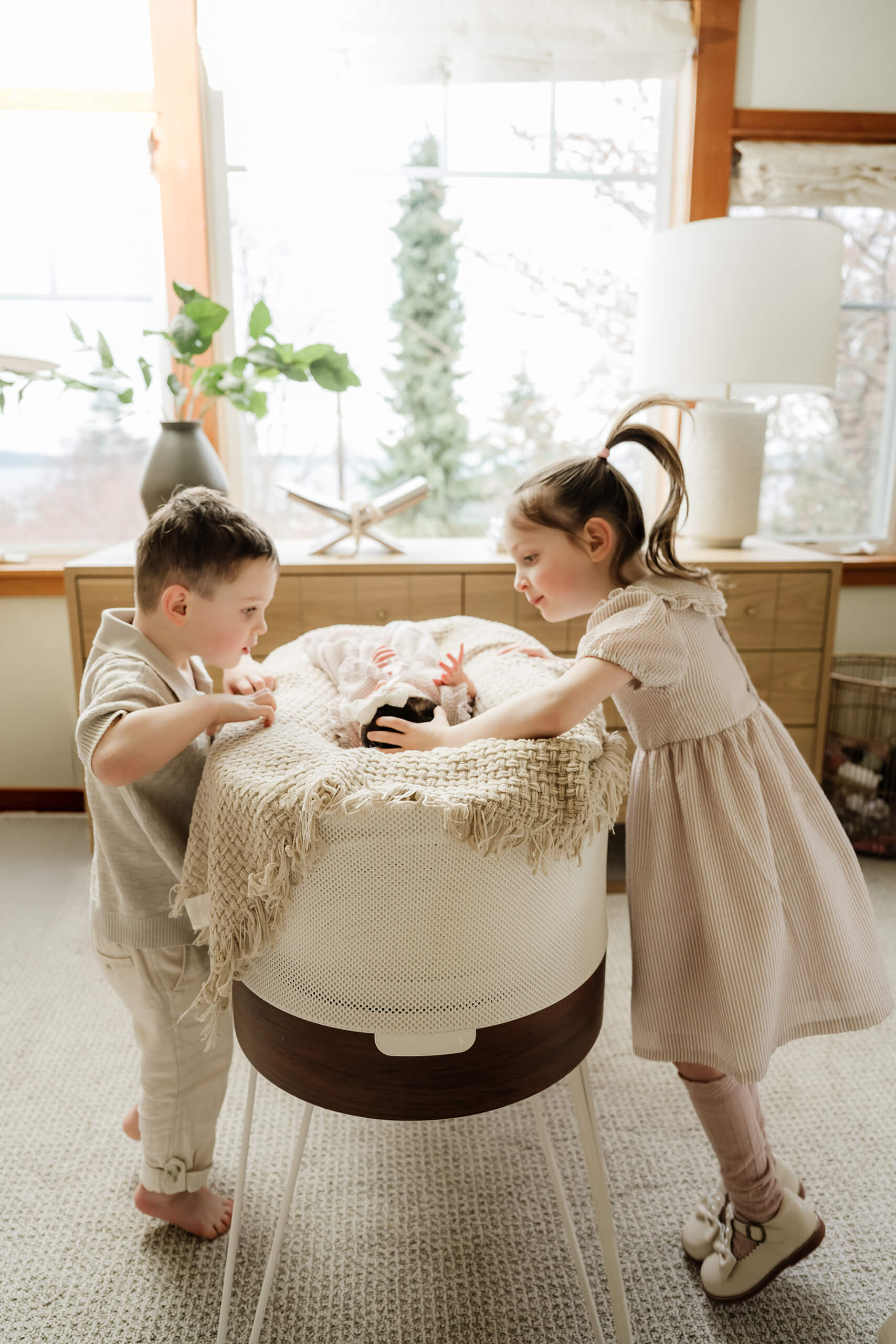 Older brother and sister stand beside the bassinet and look at their newborn sibling.