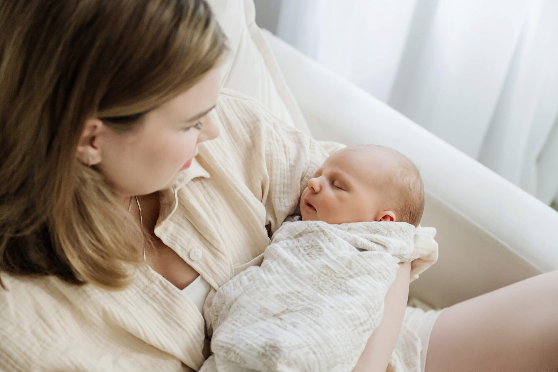 Mother looks down at her sleeping newborn baby while holding the baby near a bright window.