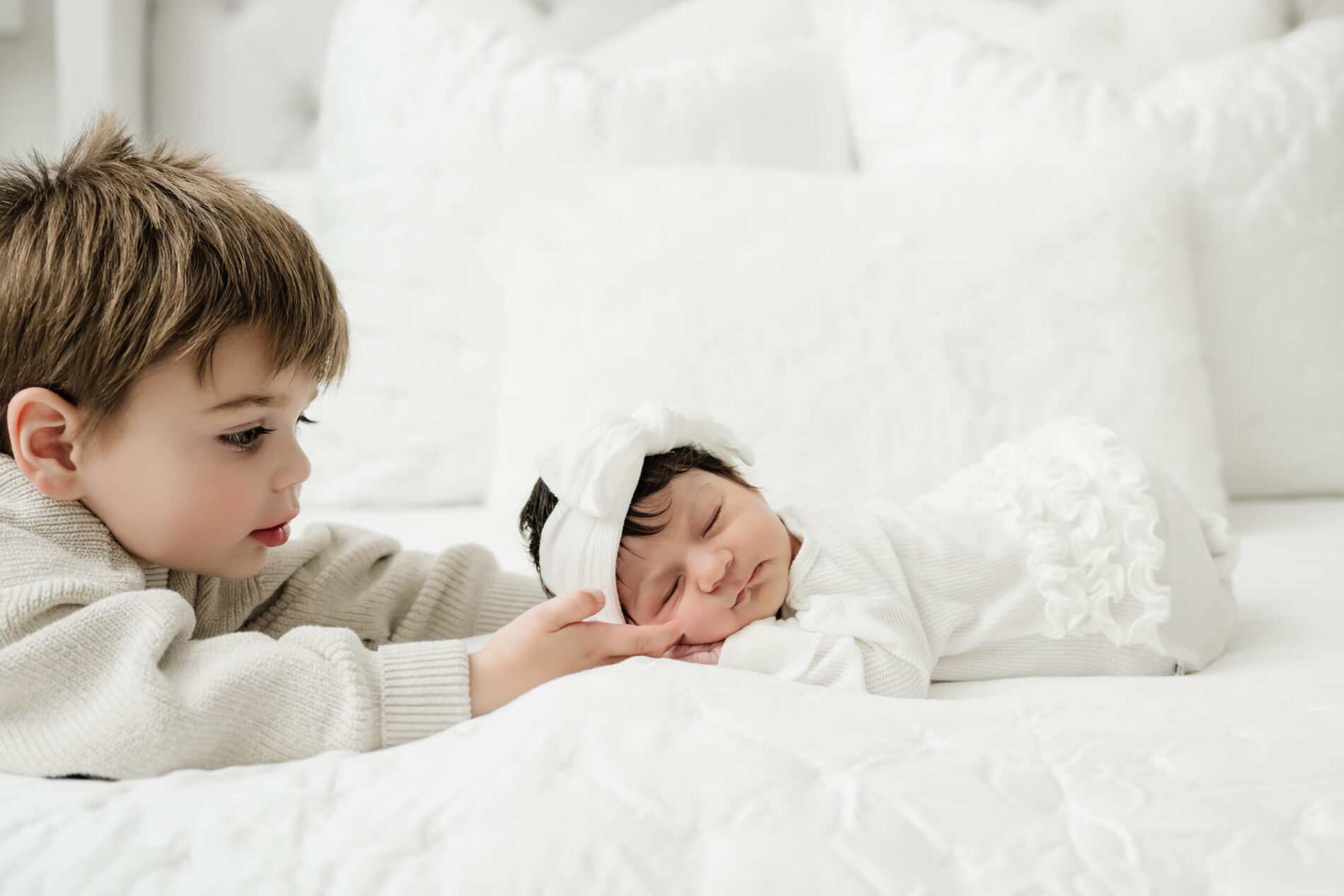 Older brother gently touches his sleeping newborn sister’s cheek as she rests on a white bed.