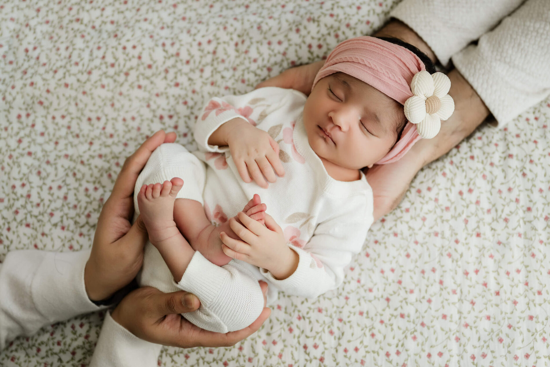 Sleeping newborn baby in a floral outfit and pink headband rests securely in a parent’s hands.
