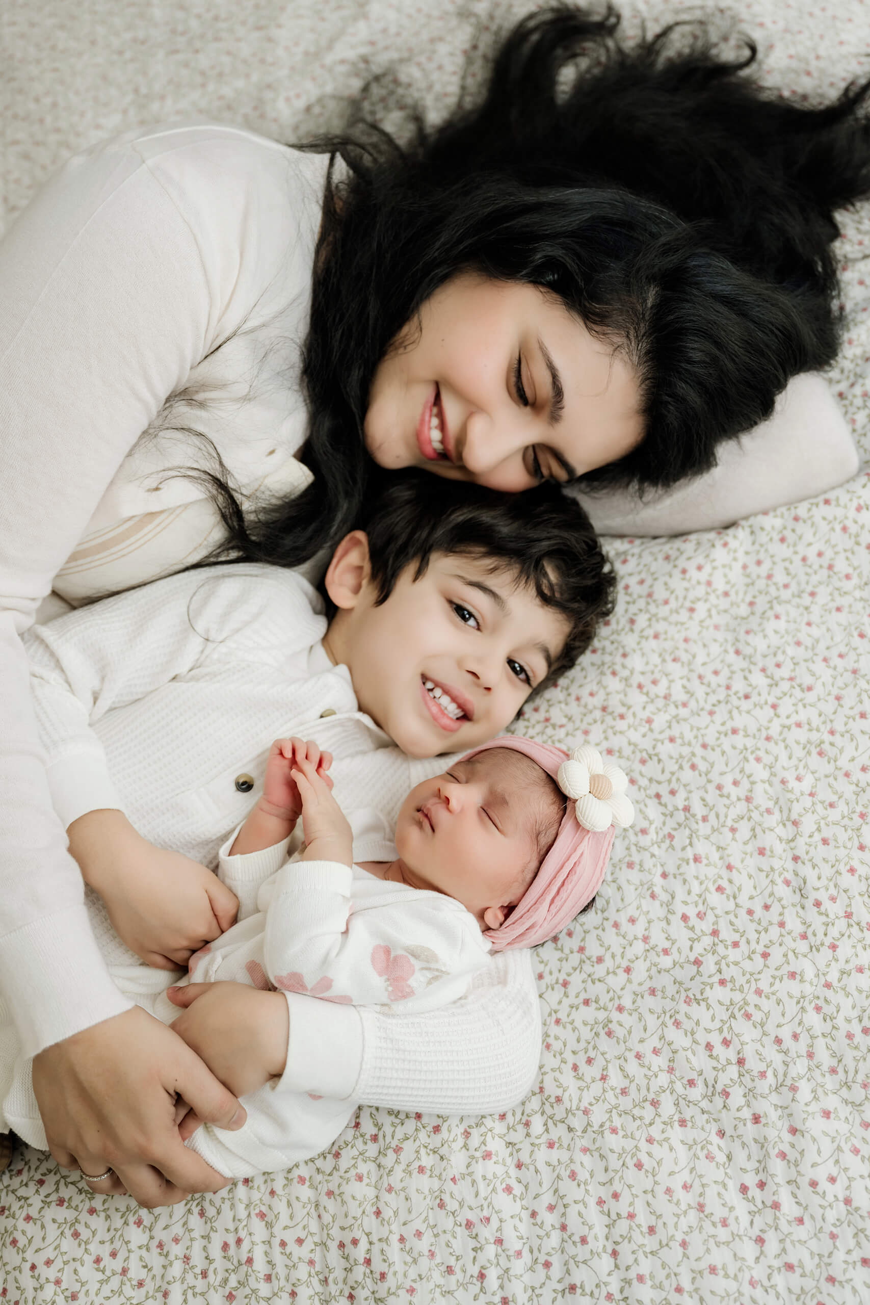 Mother, older brother, and newborn baby cuddle together on a floral bedspread.