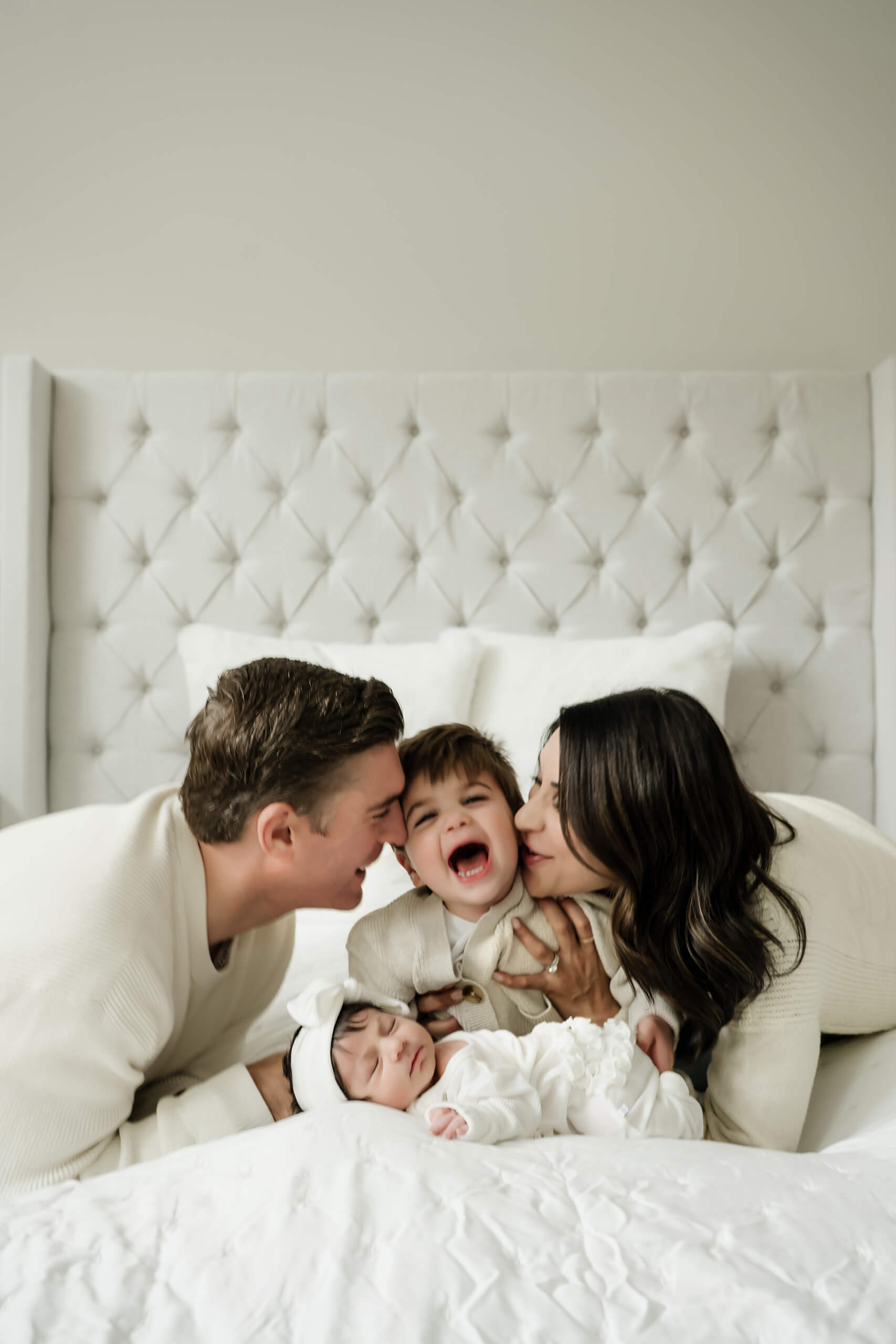 Mother and father kiss their toddler while their newborn baby sleeps beside them on the bed.