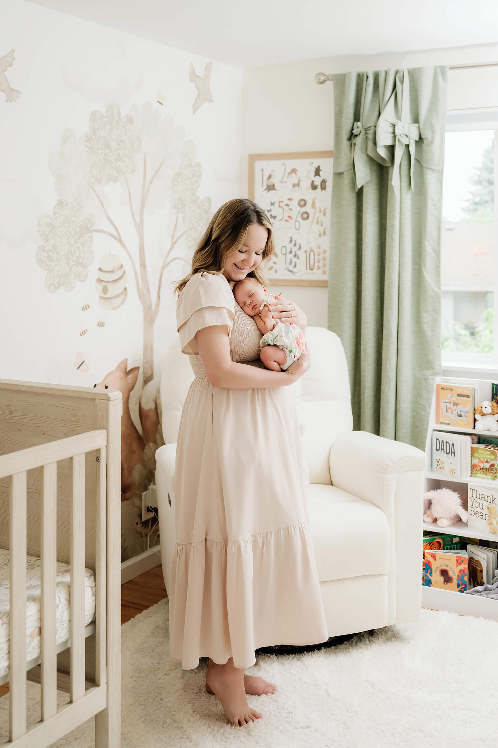 Mother stands in a woodland-themed nursery while holding her sleeping newborn baby against her chest.
