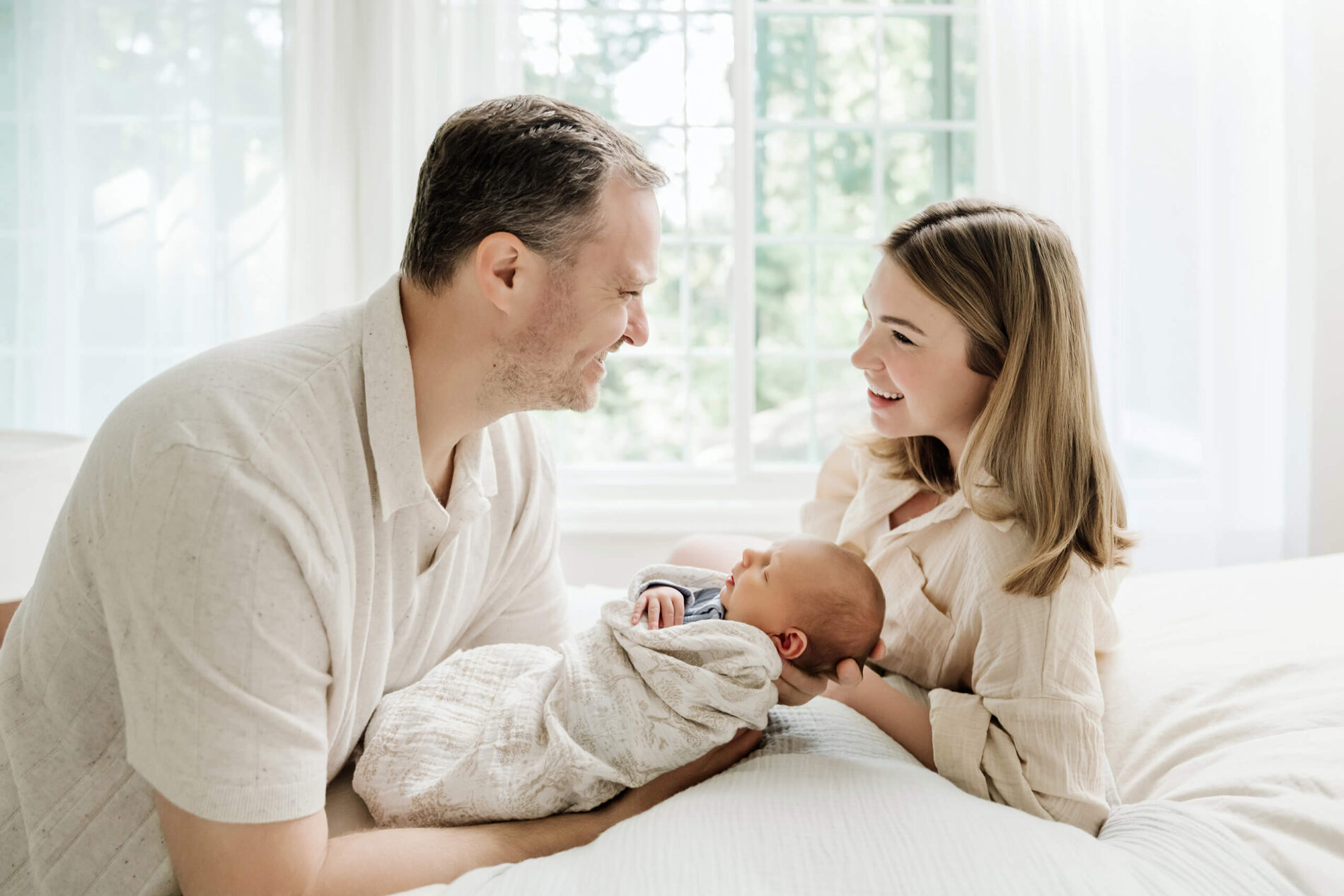 Mother and father smile at each other while cradling their newborn baby together on a bright bed.