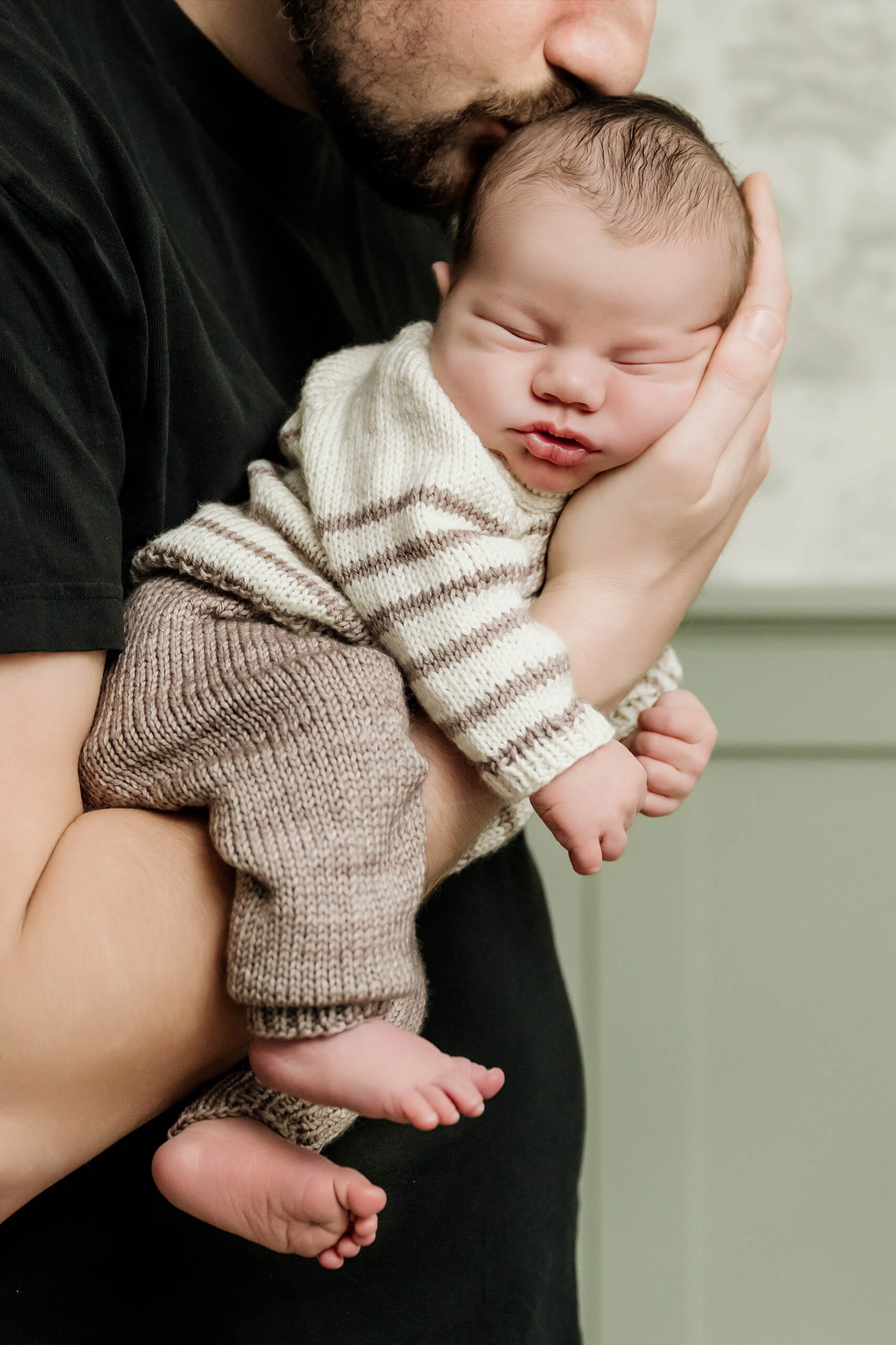 Father kisses his sleeping newborn baby’s head while holding the baby close in a knitted outfit.