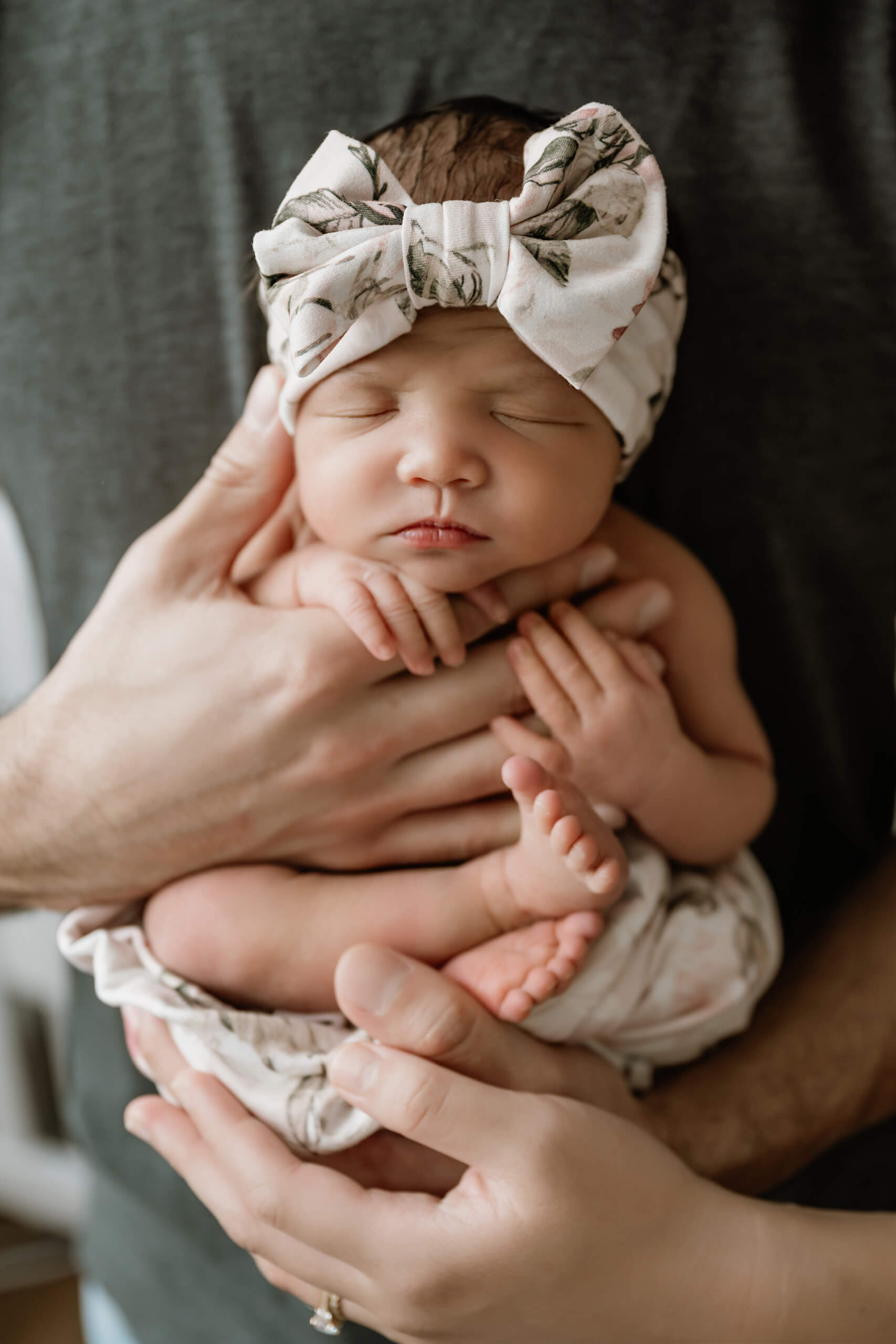Sleeping newborn girl wearing a floral bow headband is gently supported in her parents’ hands.