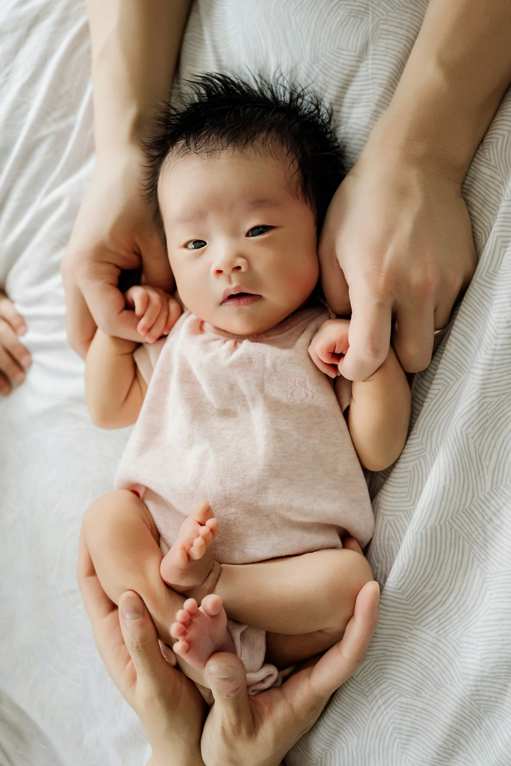 Awake newborn baby in a pale pink outfit rests securely in her parents’ hands on the bed.