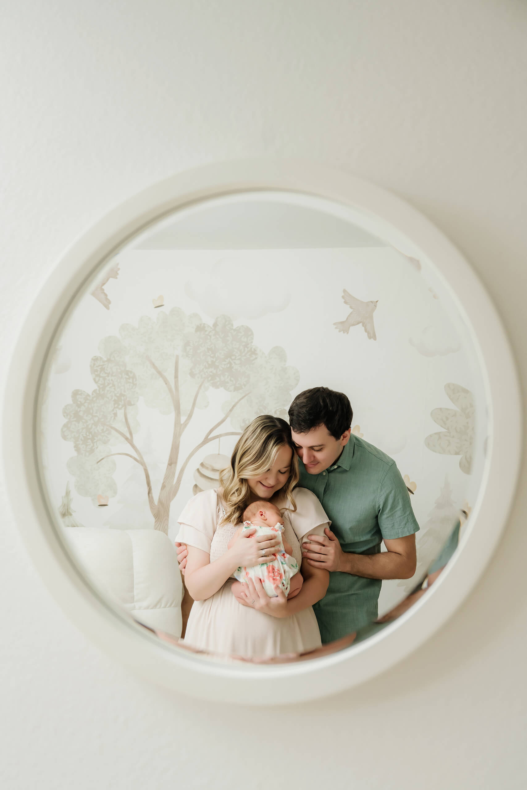 Mother and father admire their newborn baby in the reflection of a round nursery mirror.