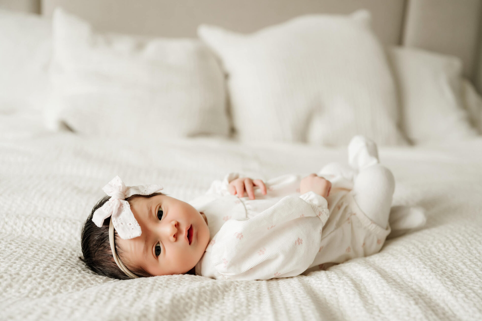 Awake newborn girl wearing a white outfit and bow headband lies comfortably on a white bed.