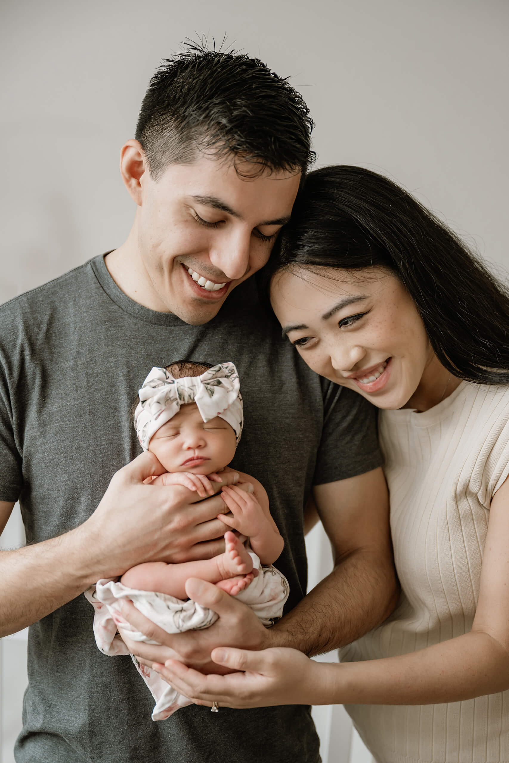 Mother and father smile down at their sleeping newborn daughter while gently holding her together.
