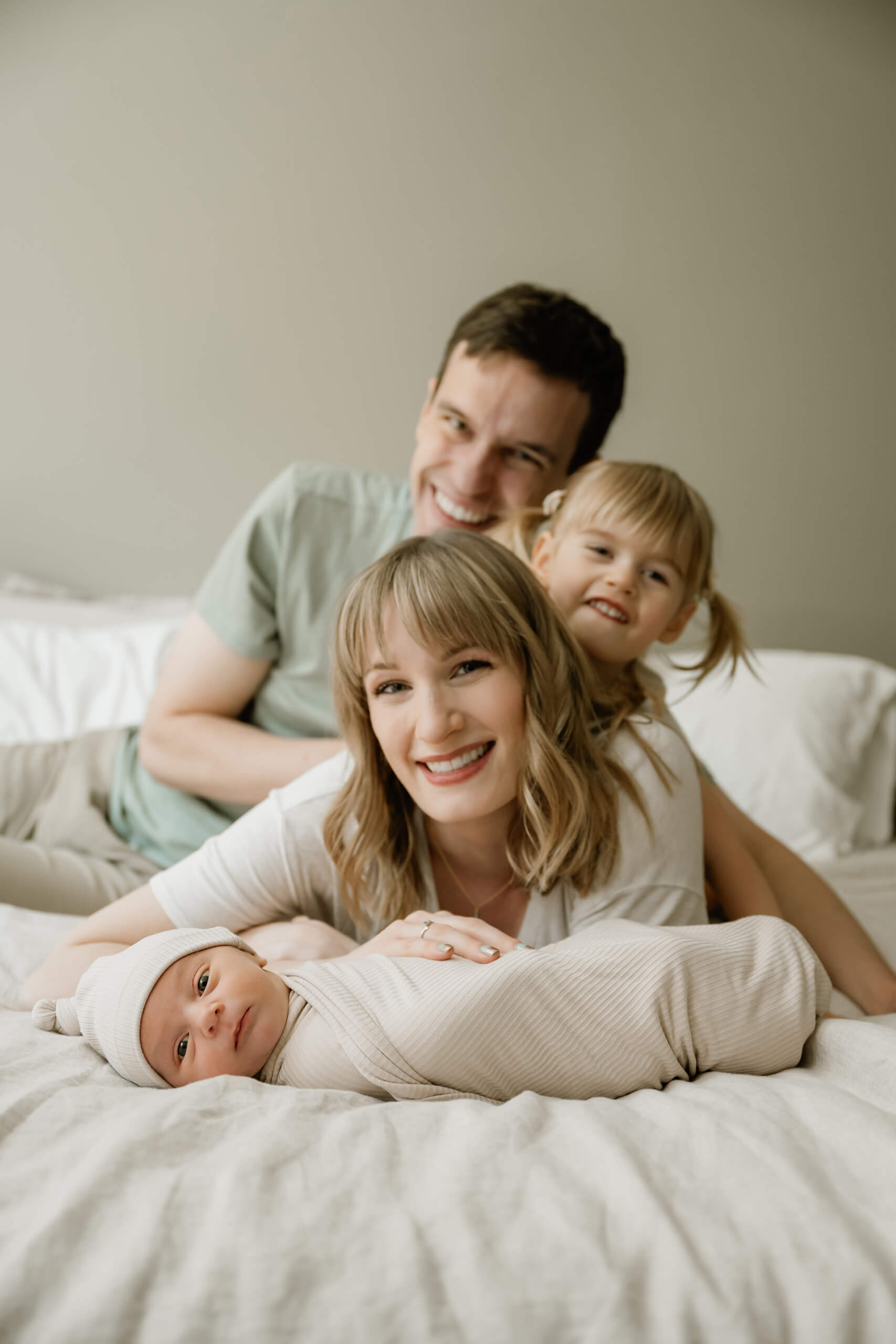 Family of four gathers on a bed behind their awake newborn baby wrapped in a neutral striped blanket.