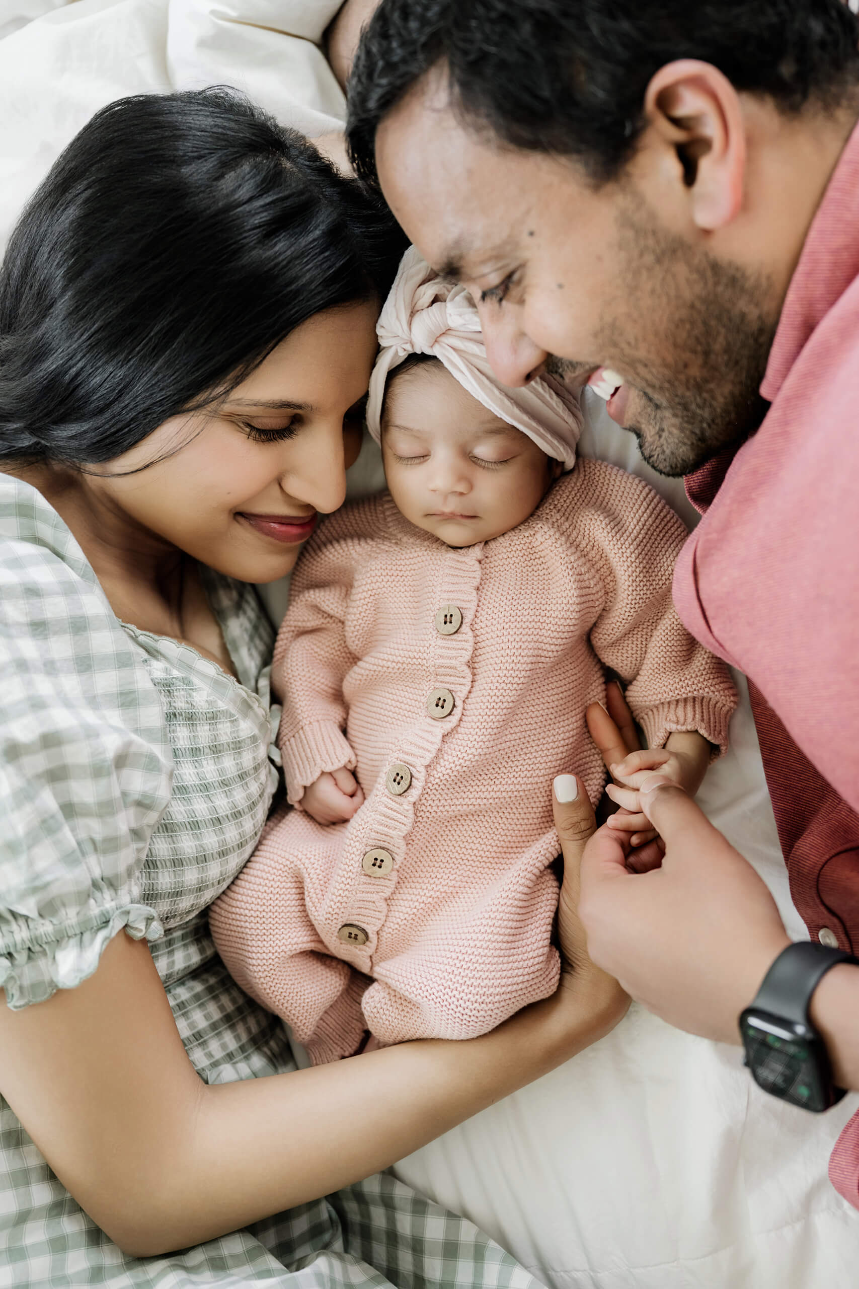 Mother and father lie beside their sleeping newborn daughter dressed in a soft pink knitted outfit.