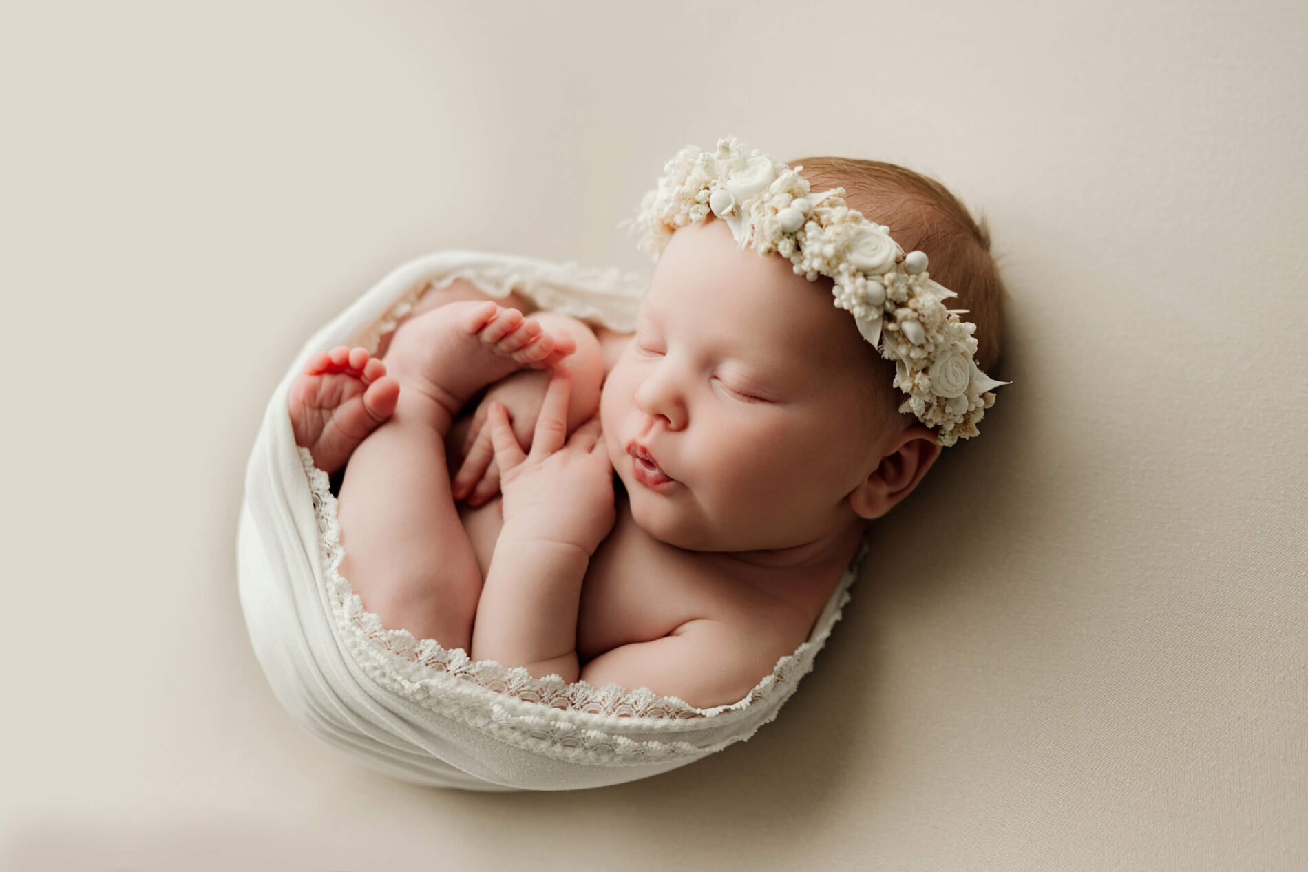 Newborn with eyes closed curls inside a white wrap while wearing a cream floral headband.