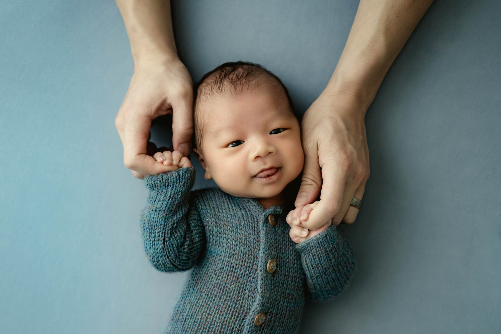 Awake newborn in a teal knit outfit lies on a blue backdrop while an adult holds both hands.