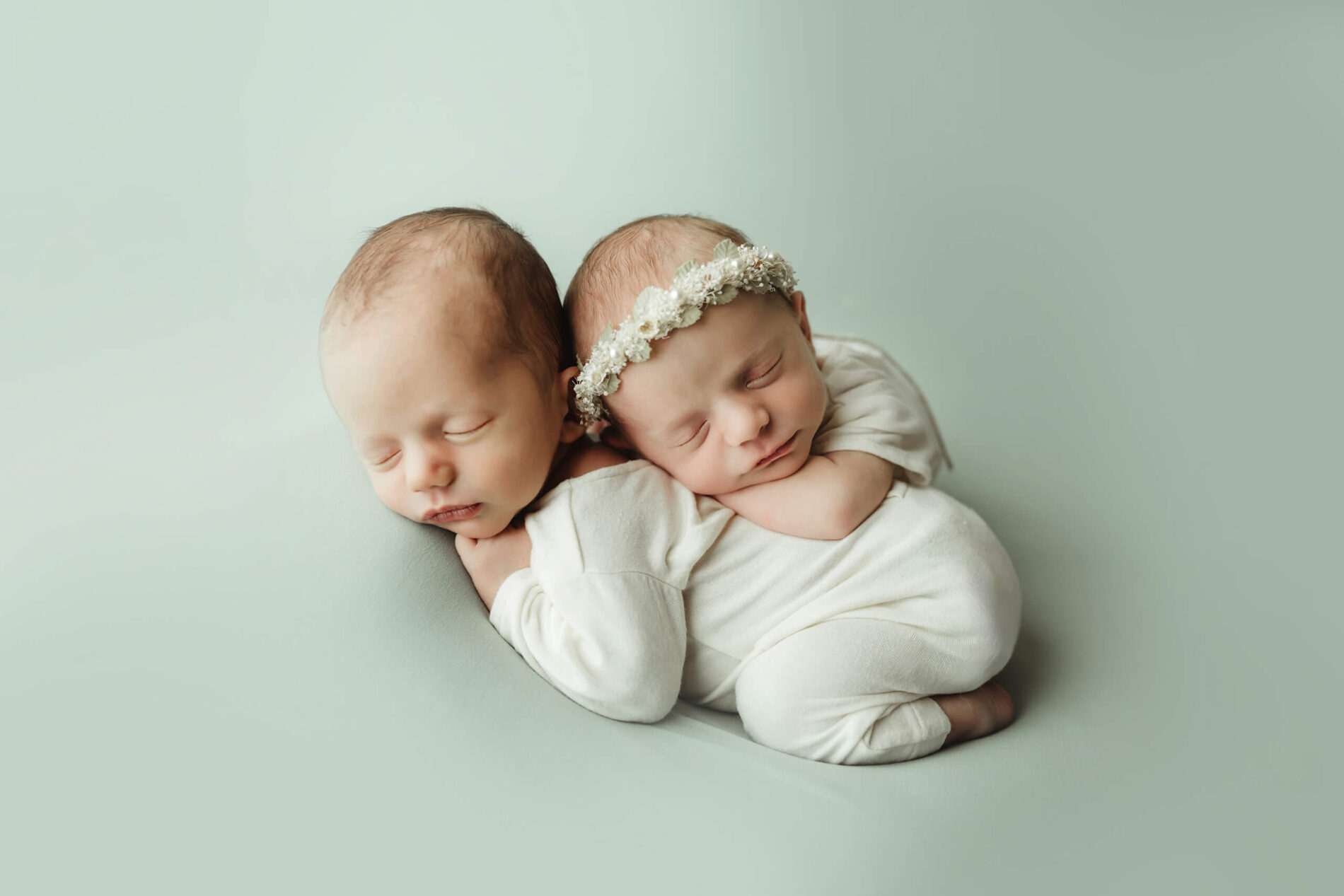 Two newborns with eyes closed rest together in white outfits, one wearing a cream floral headband.