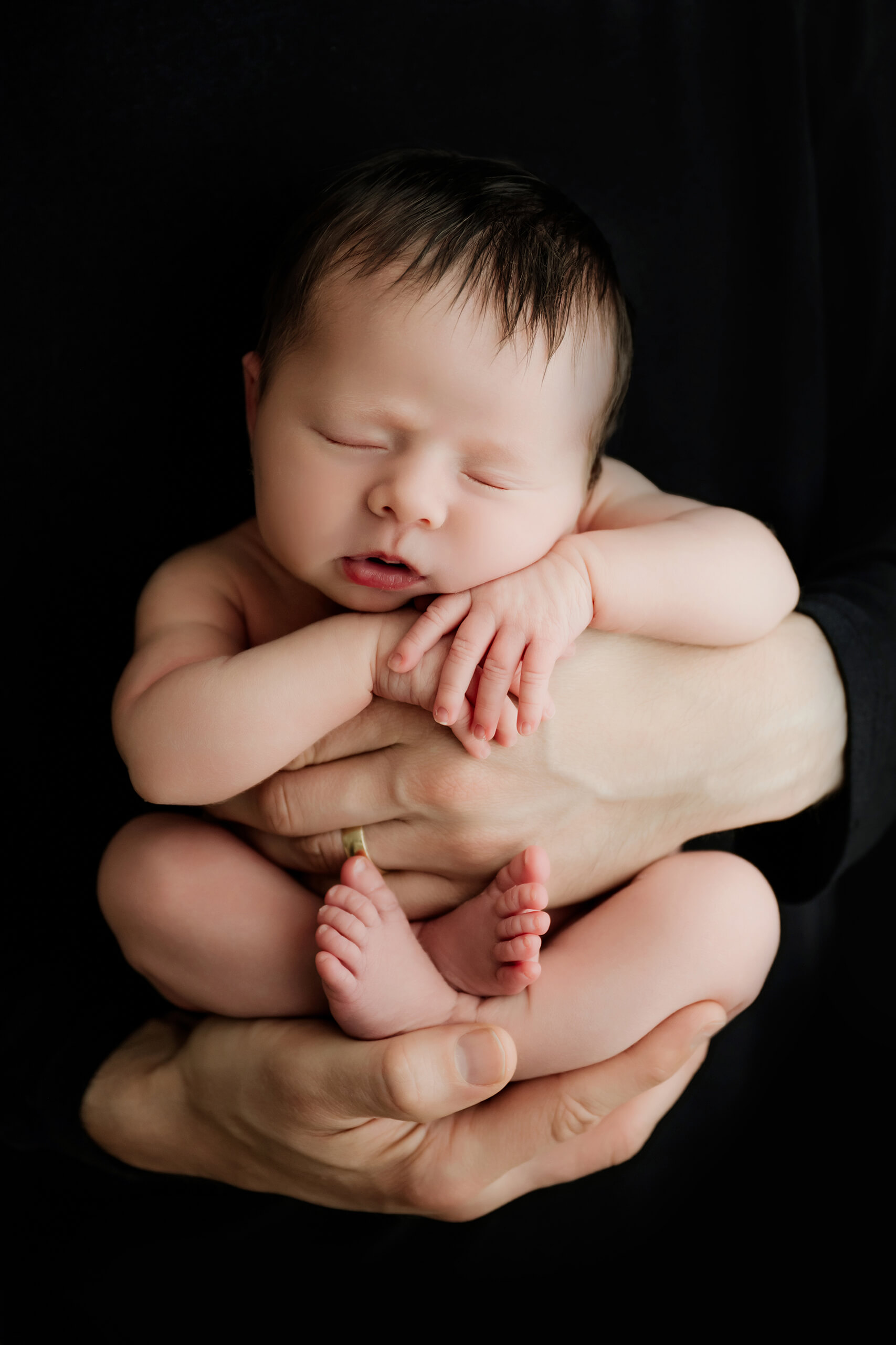 Unwrapped newborn with eyes closed is supported upright in adult hands against a black background.