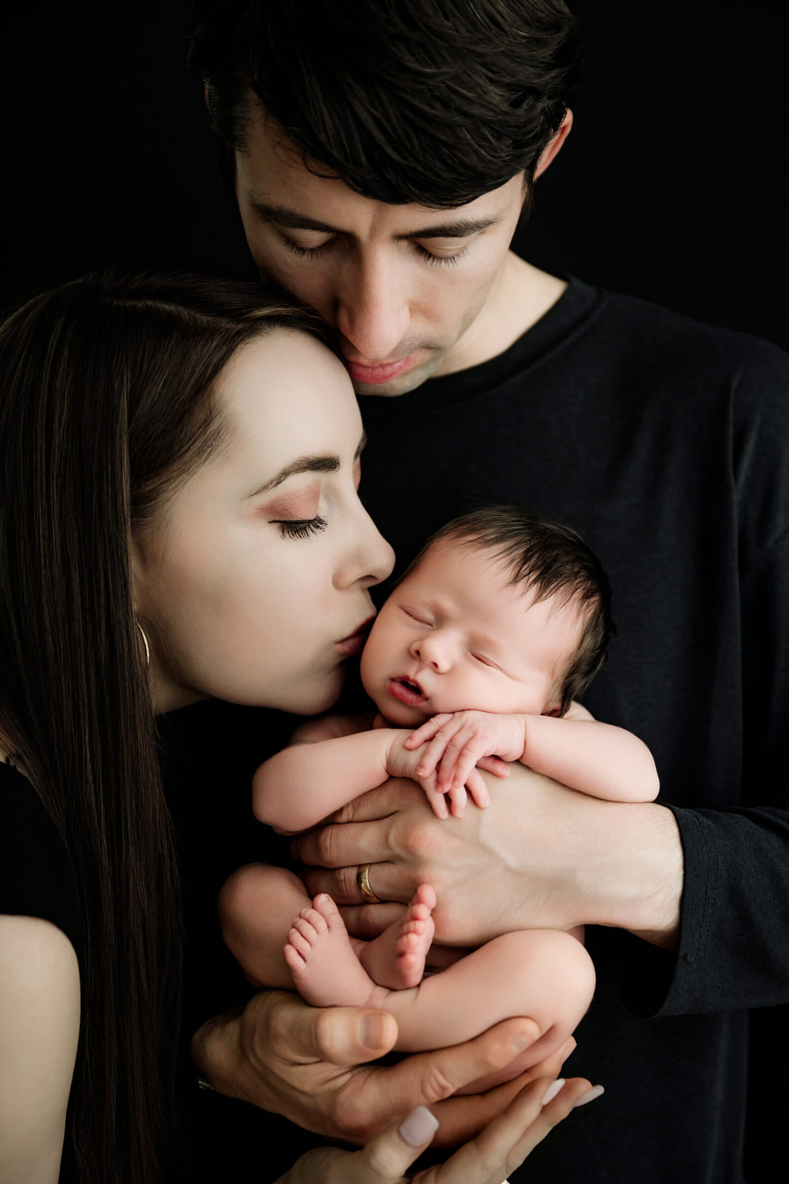 Two parents hold an unwrapped newborn as one kisses the baby’s cheek against a black background.