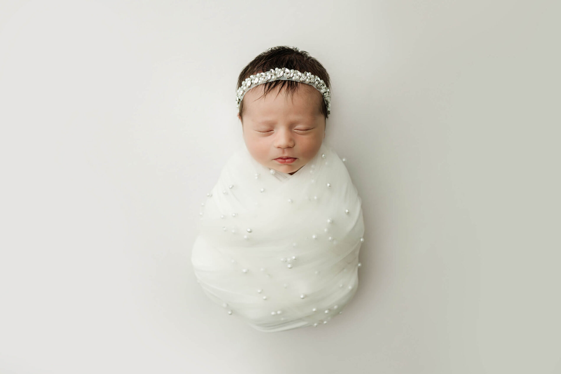 White-wrapped newborn with eyes closed wears a jeweled headband against a simple white background.